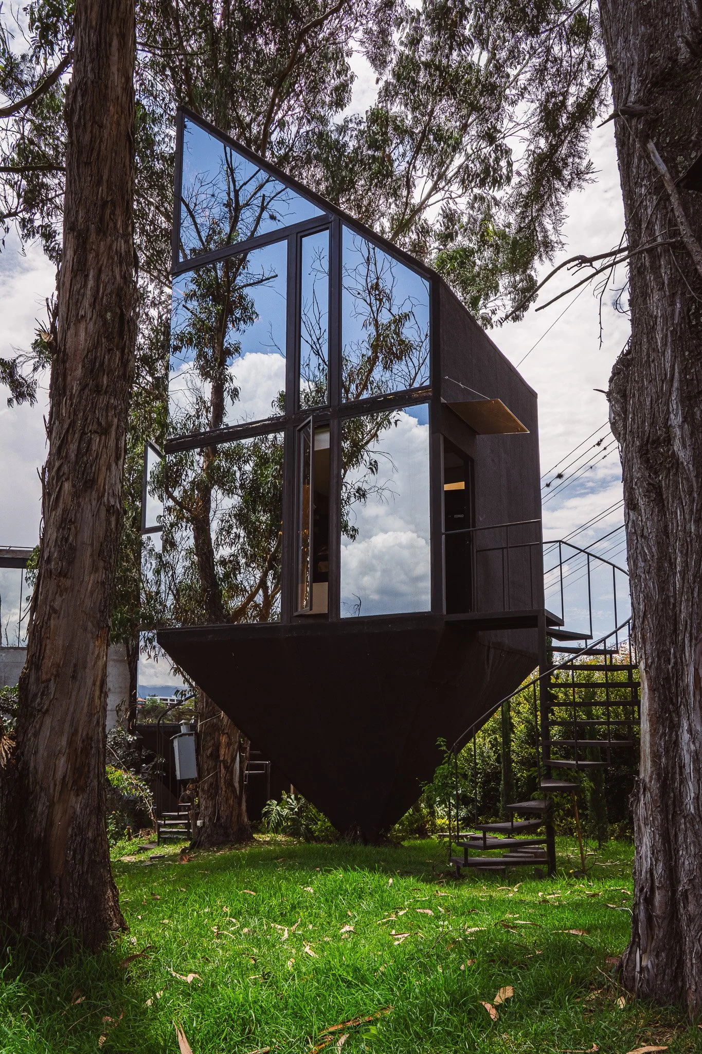 A modern black house with large reflective glass windows is built on stilts among tall trees with textured bark. The background shows a cloudy sky and surrounding trees, with a small staircase leading up to the house and lush green grass beneath.