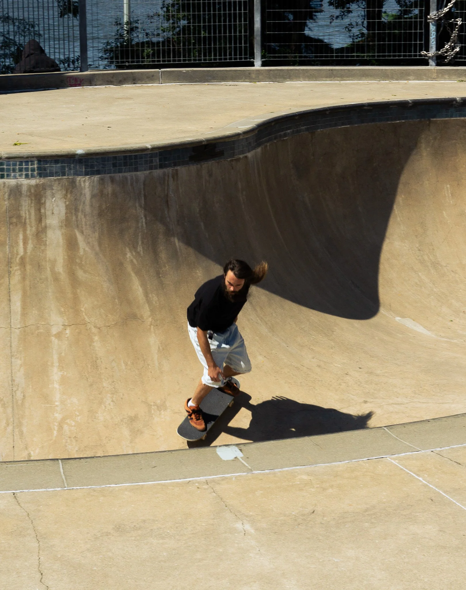 Teenage boy skateboarding in a skatepark, seen through a mirror or reflective surface, with a fence and water in the background.