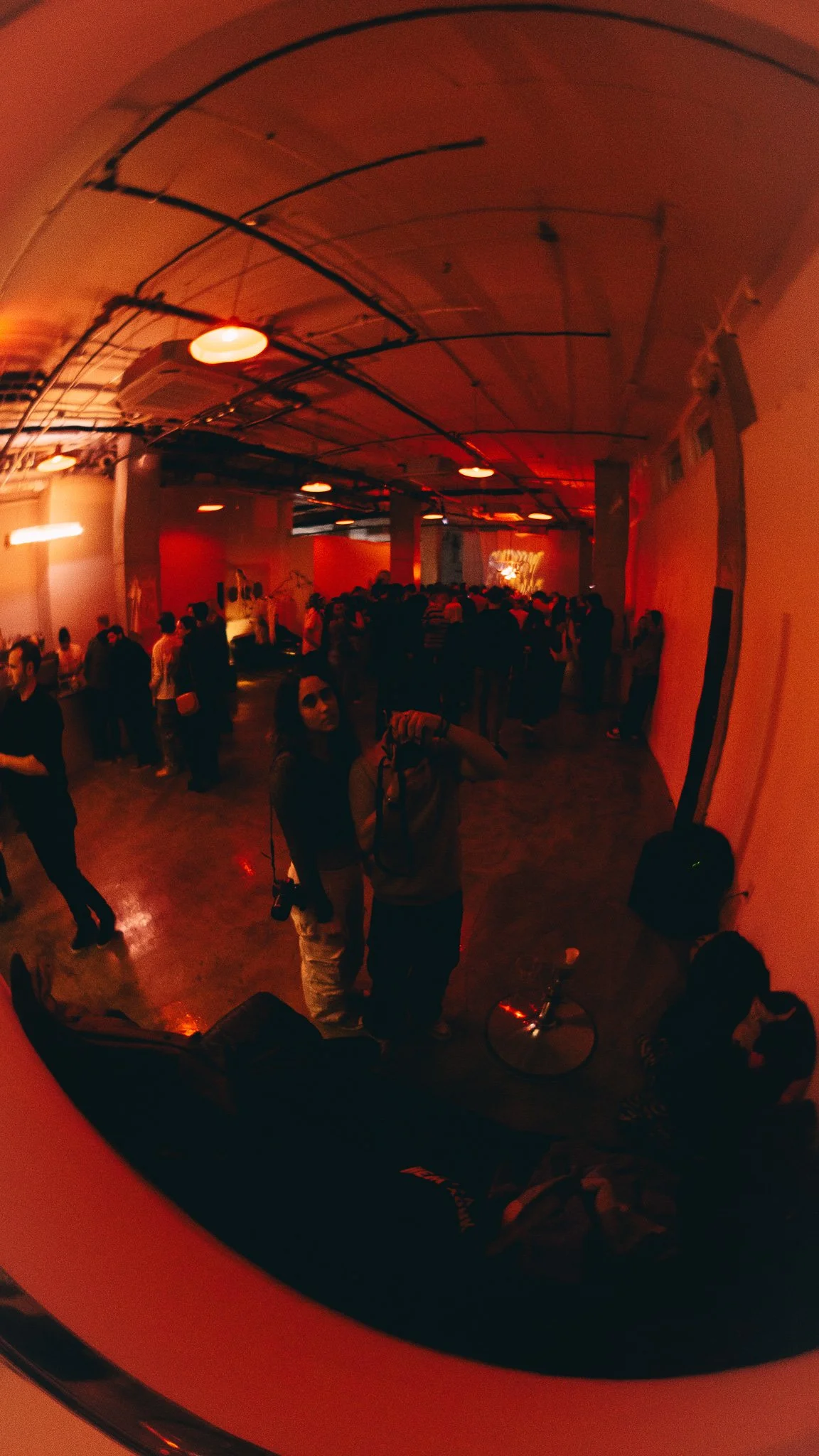People gathered in an indoor venue with red lighting and a grungy industrial style ceiling, some of them are standing and talking, with a woman wearing sunglasses in the foreground.