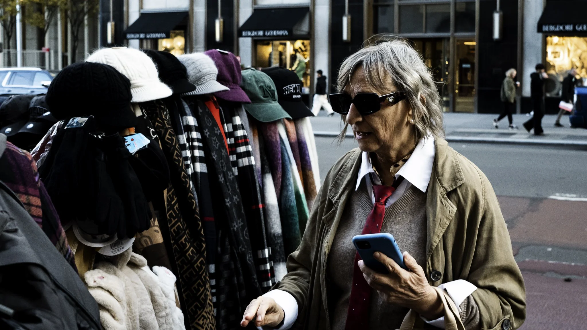An elderly woman with gray hair, wearing black sunglasses, a beige trench coat, white shirt, pink tie, and holding a blue smartphone, stands on a city sidewalk across from a street vendor stall displaying various colorful scarves and hats. In the bac