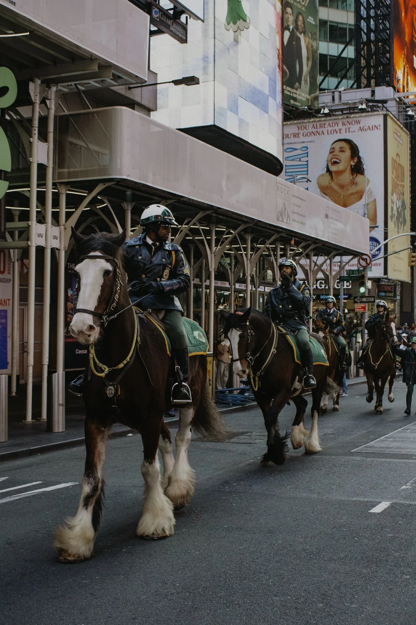City street scene with mounted police officers on large horses, surrounded by tall buildings with digital billboards and advertisements, including a smiling woman and colorful signs.
