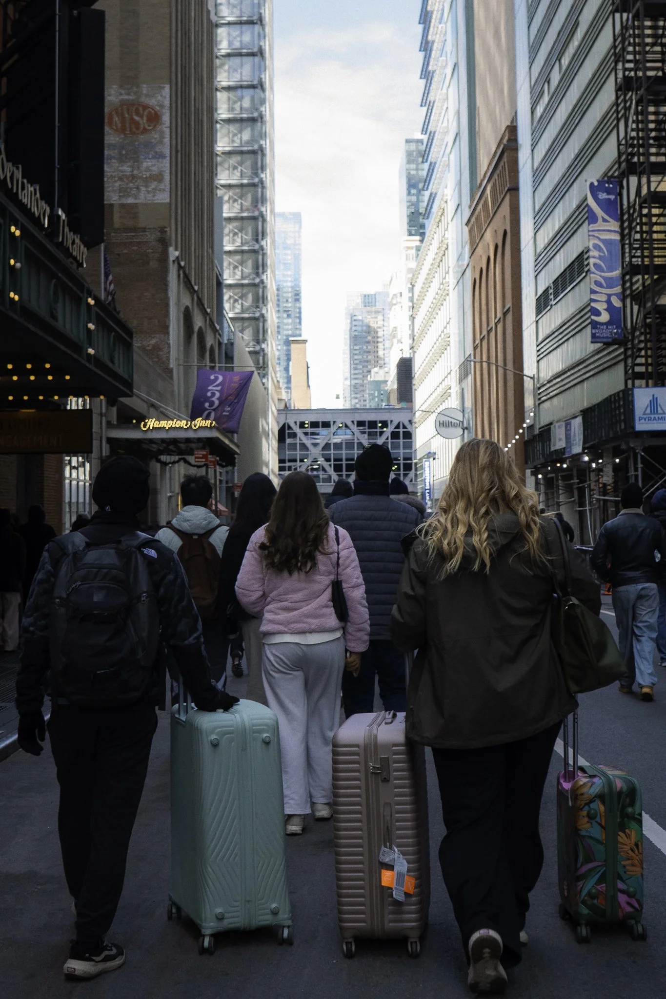 Group of people walking through city street pulling rolling luggage, buildings and signs visible in the background.