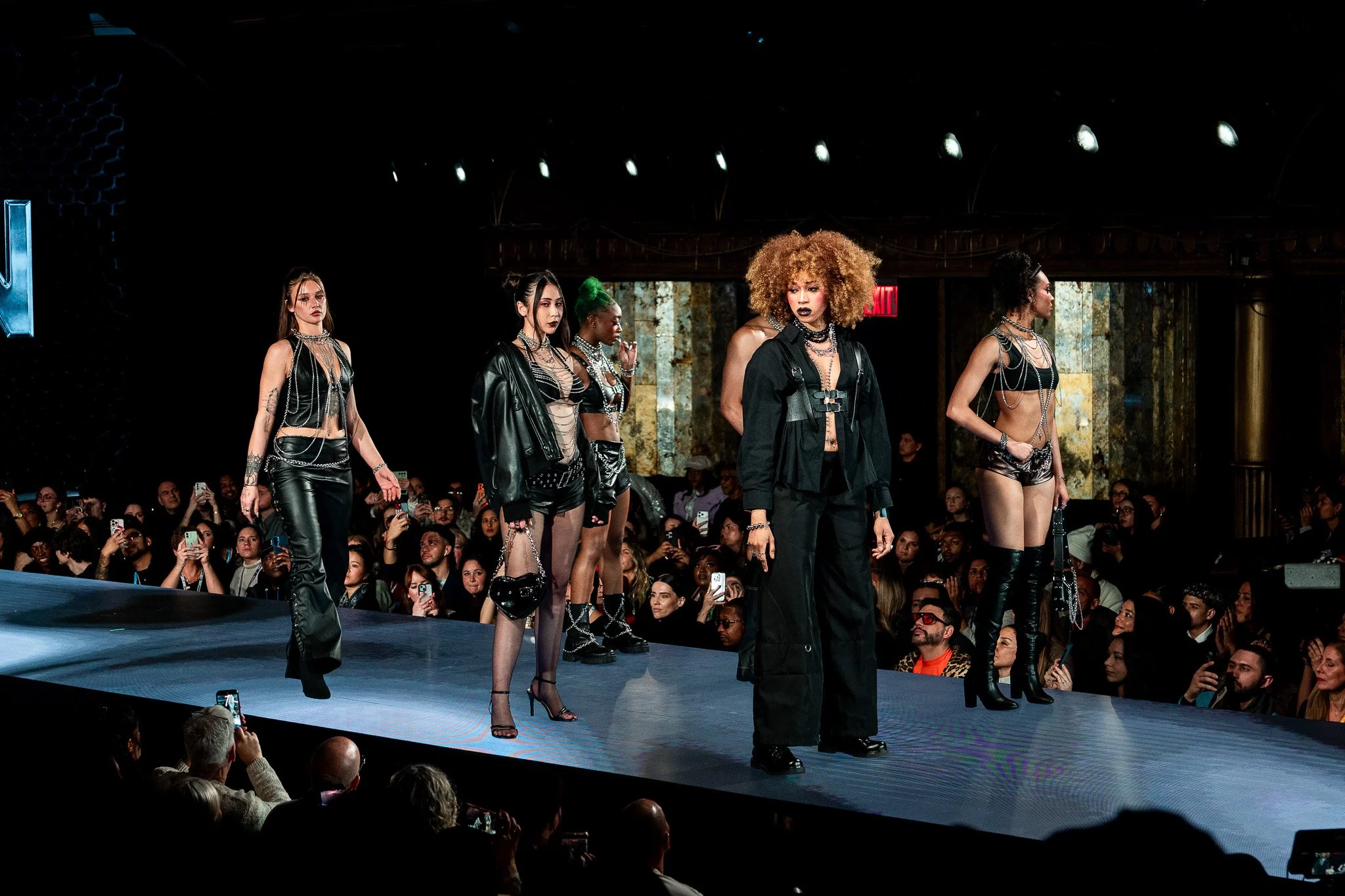 Fashion models walking on runway during a fashion show with audience taking photos.