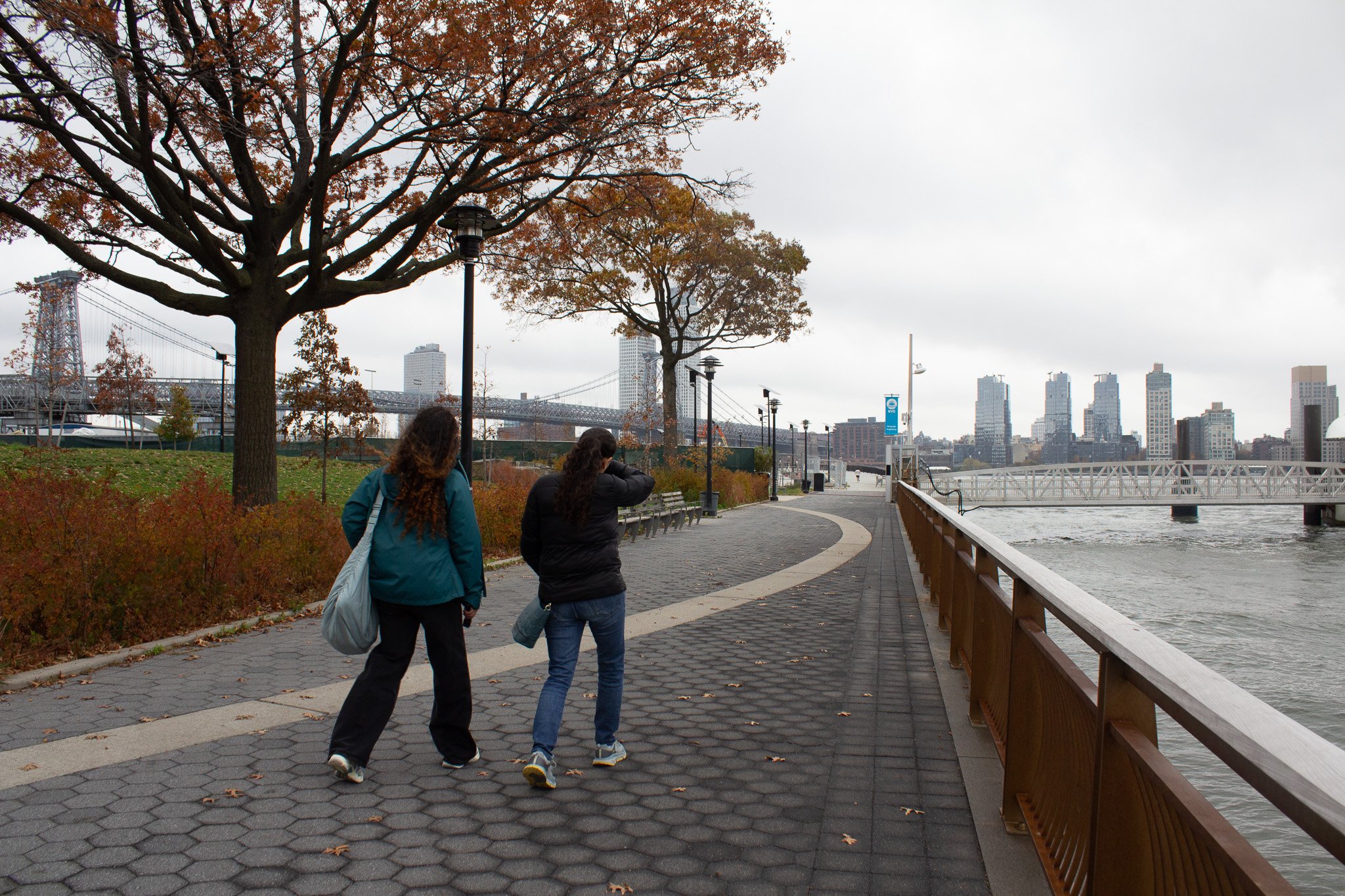 Two women walking along a riverside path in an urban park under autumn trees with a city skyline and bridge in the background on a cloudy day.