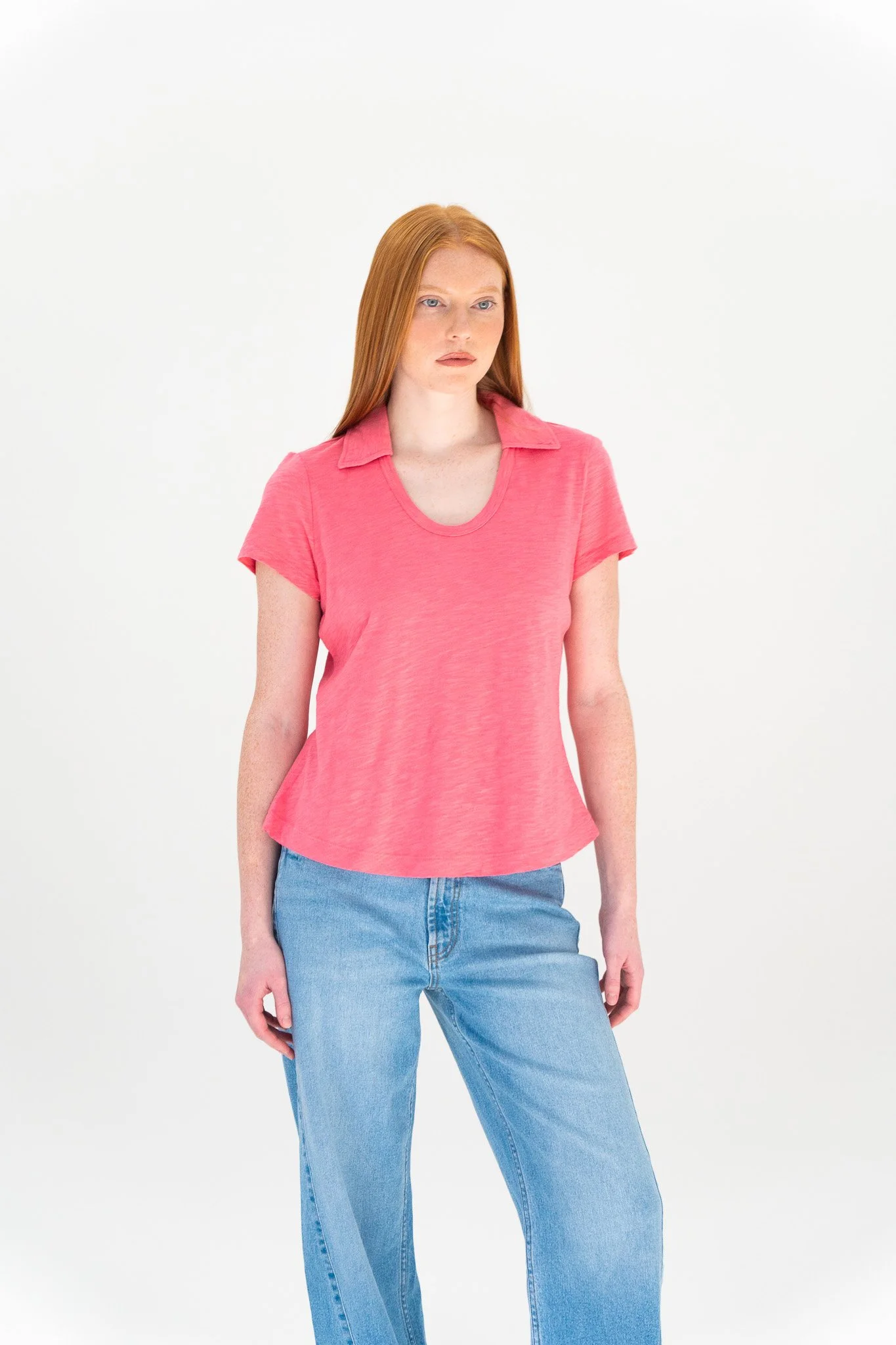 A woman with long red hair, wearing a pink T-shirt and light blue jeans, standing against a plain white background.