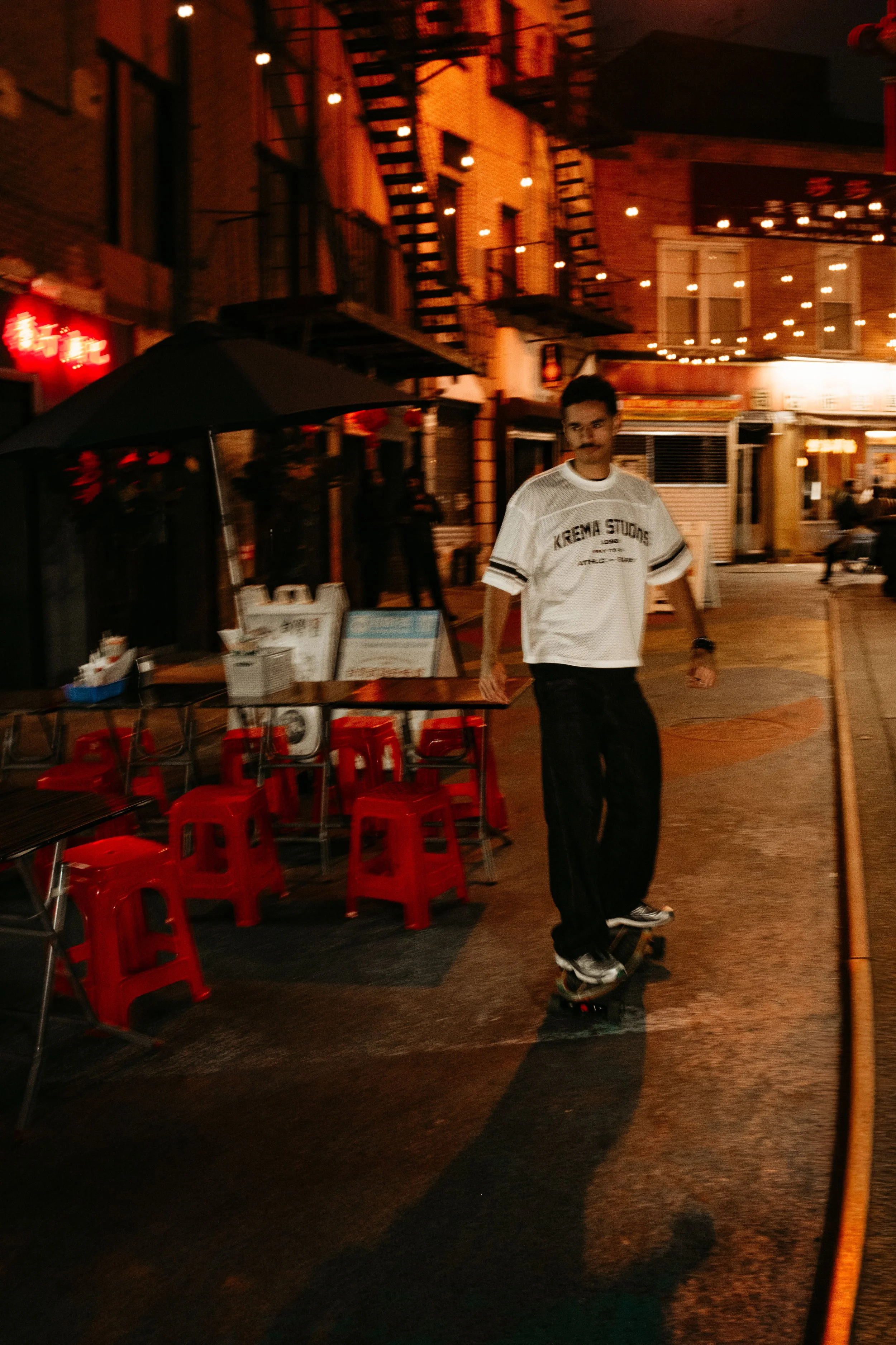 A young man skateboarding on a city street at night, illuminated by warm string lights, with outdoor seating and red stools nearby.