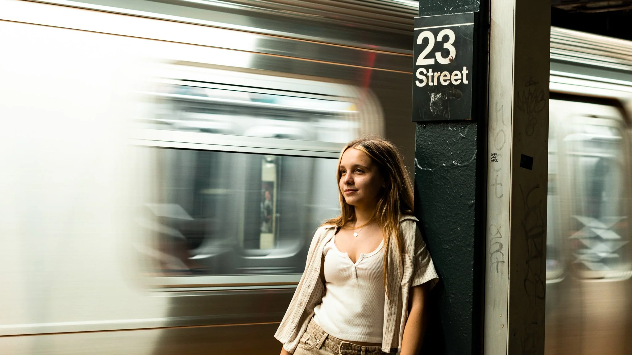 A young woman with long brown hair, wearing a white shirt and beige pants, standing on a city subway platform next to a black pillar with the sign '23 Street.' A train is passing by at high speed behind her.