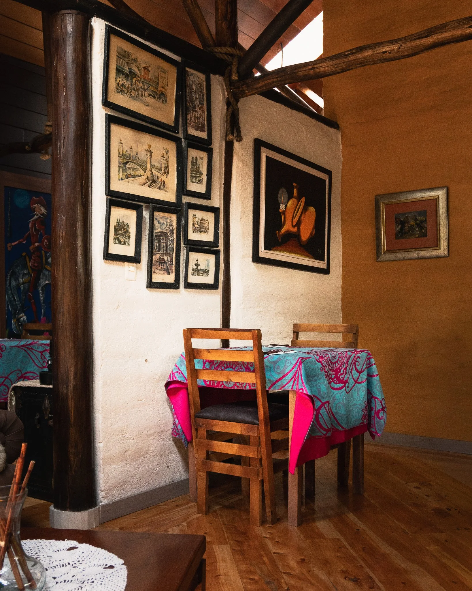 A cozy corner of a room with a wooden table covered by a colorful tablecloth and a wooden chair. On the wall, there are framed pictures, including paintings of city scenes and a modern still life. Part of a wooden beam and columns are visible, with s