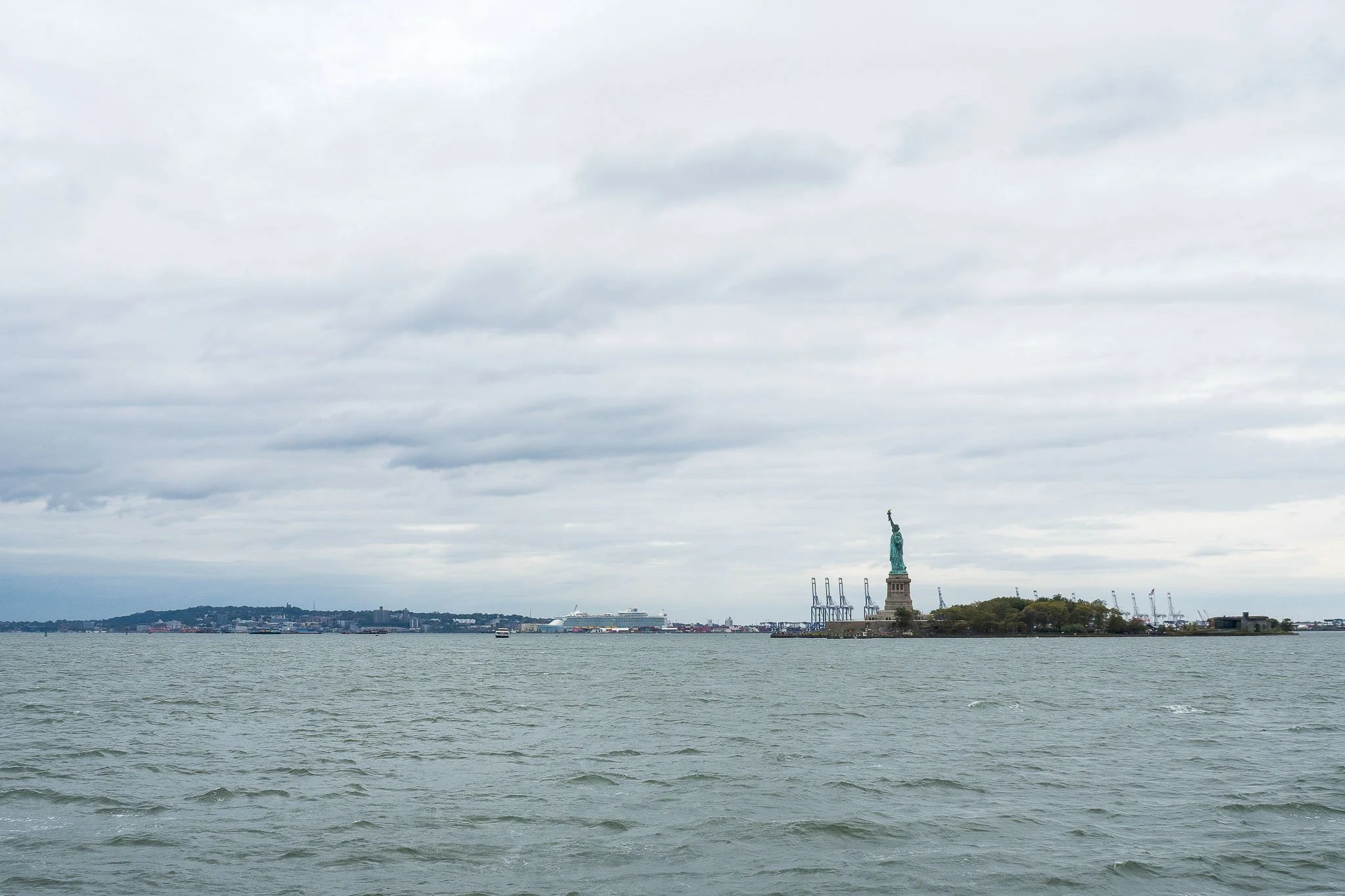 The Statue of Liberty on Liberty Island in New York Harbor, with a cloudy sky overhead and cargo ships docked at the port in the background.