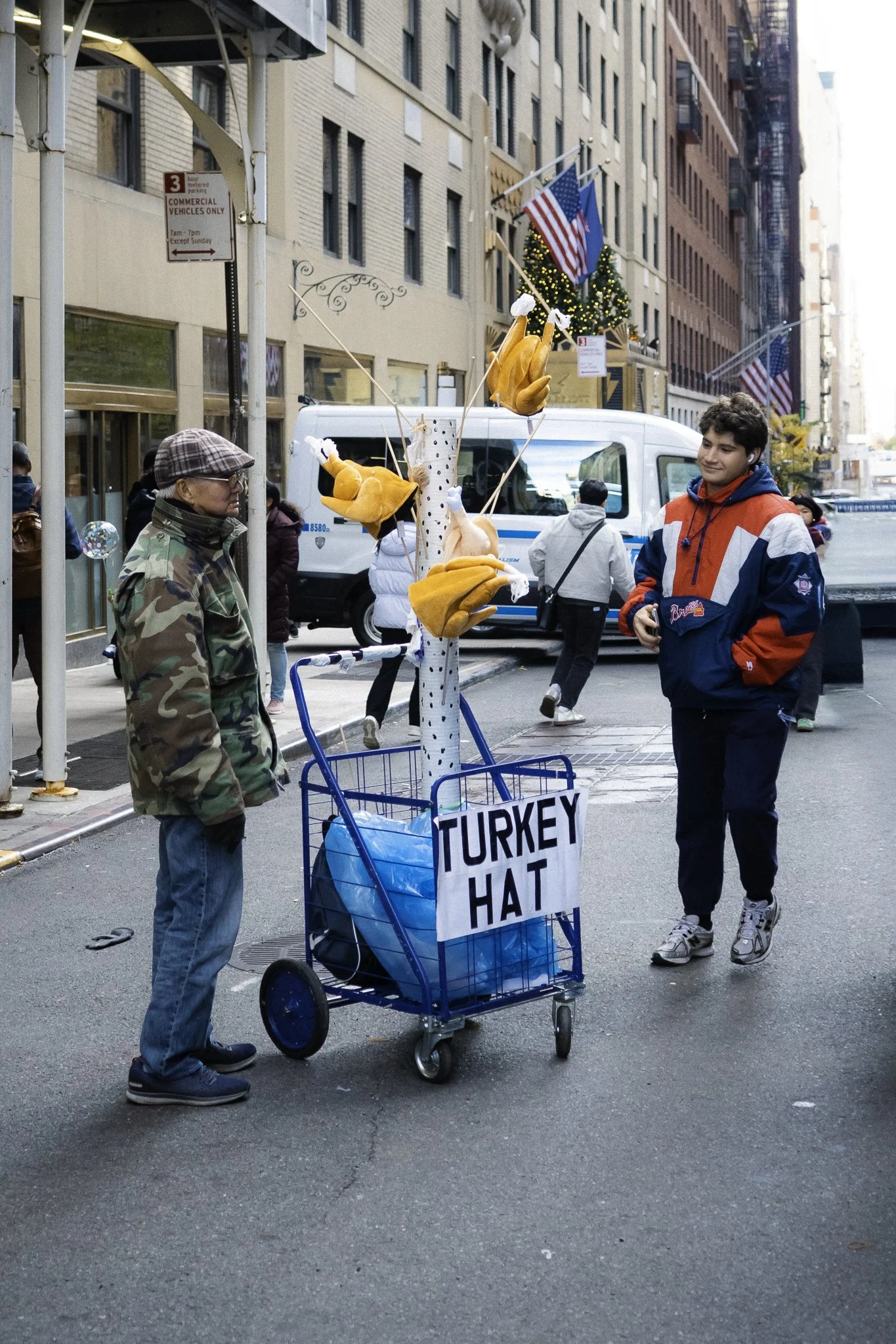 Street scene showing a man with glasses and a plaid cap standing near a shopping cart labeled 'TURKEY HAT', with a woman in a red and blue jacket walking past. The cart has plush turkey hats attached. In the background, there are buses, buildings, fl