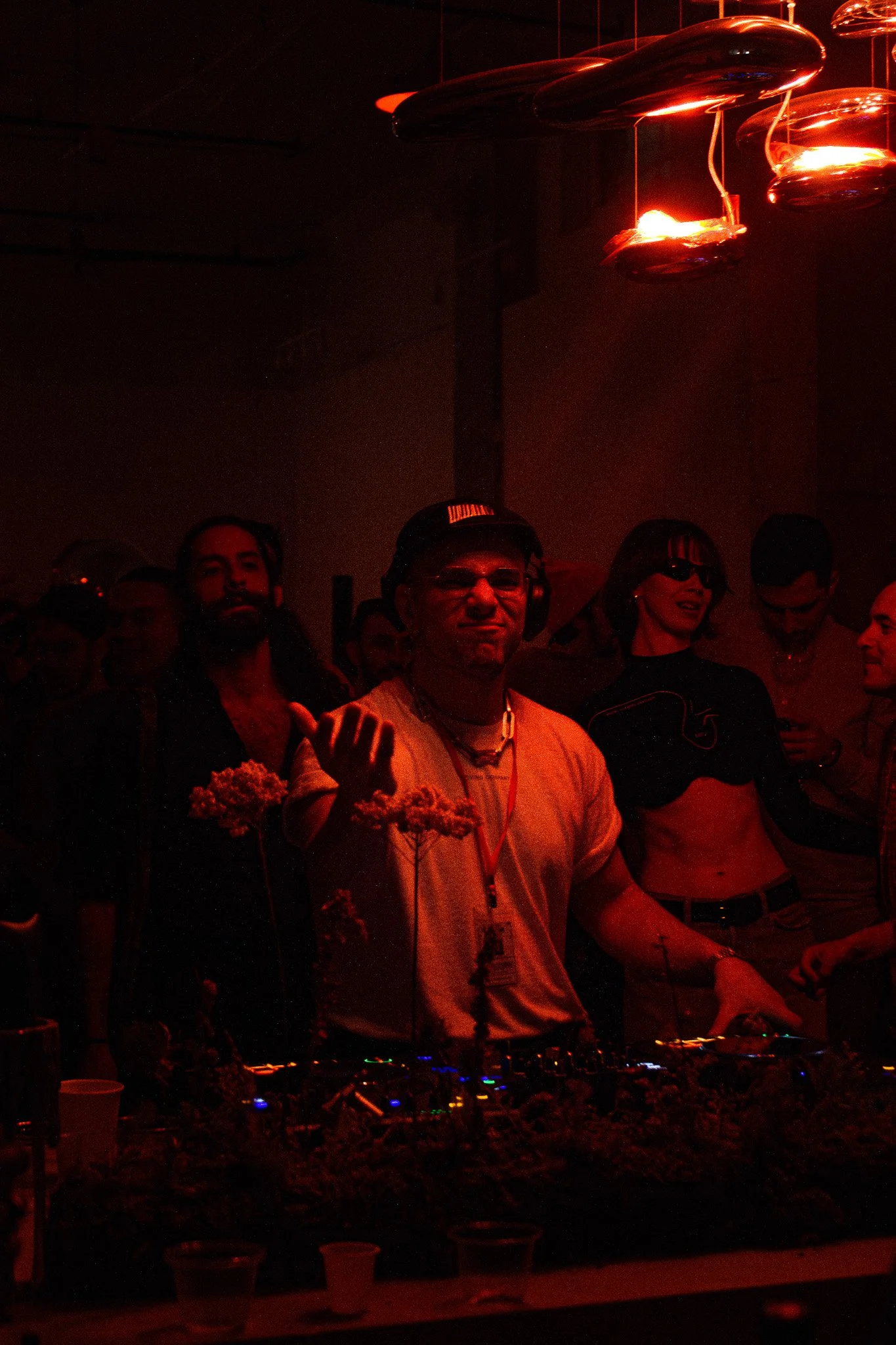 People at a nightclub or music event with dark lighting and red glow, some wearing headphones, standing behind a DJ setup.