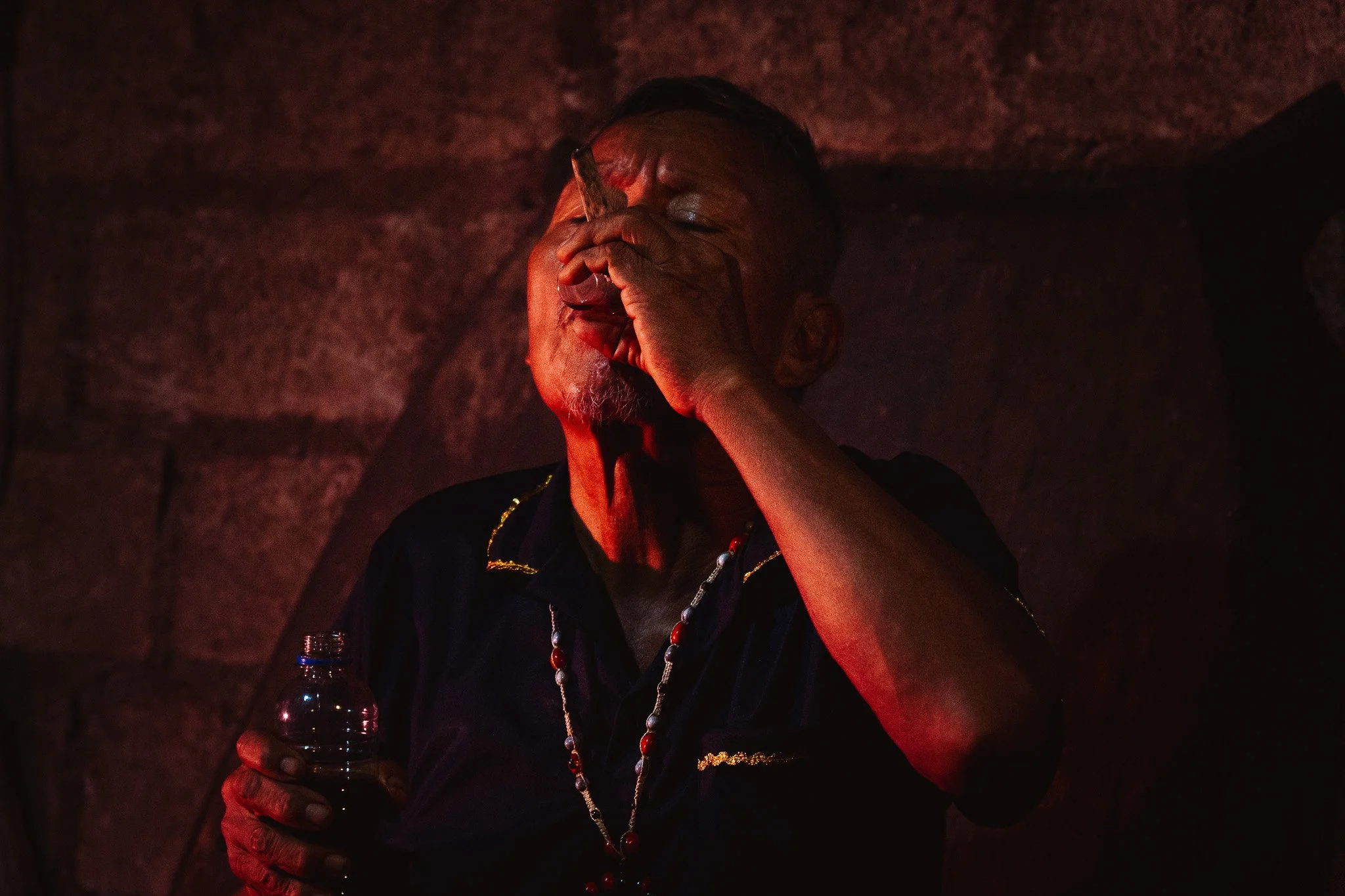An elderly person with dark hair and tan skin, covering their face with their hand, holding a water bottle, in a dimly lit setting with reddish and dark tones.