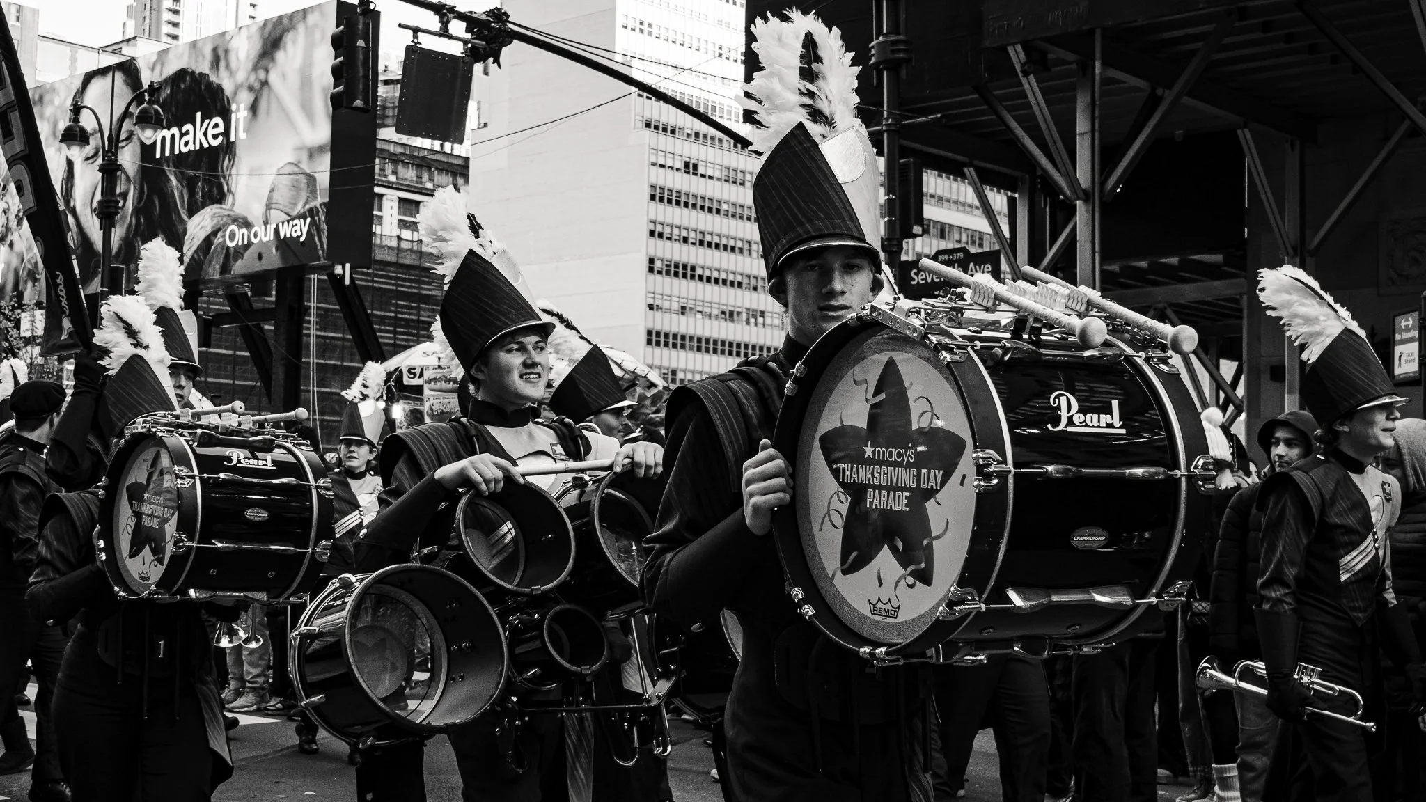 A group of marching band members playing drums during a parade in an urban area, with tall buildings and signs visible in the background.