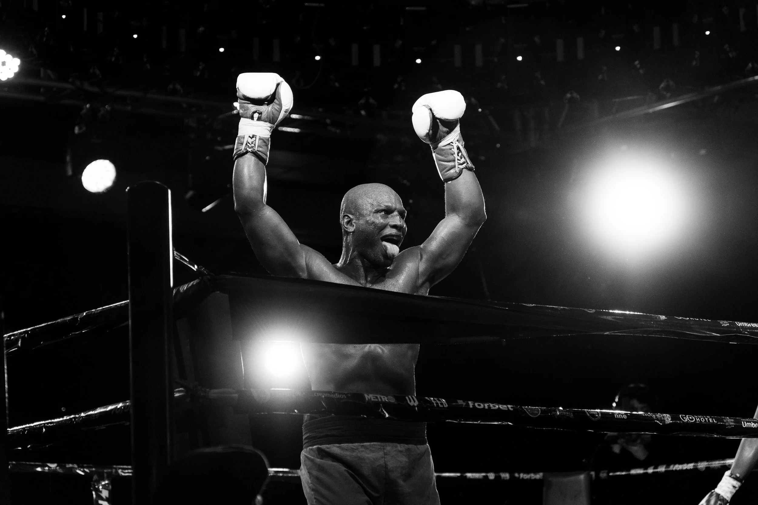 A boxer stands in the ring with both fists raised, wearing boxing gloves. The photo is black and white, capturing his triumphant expression.