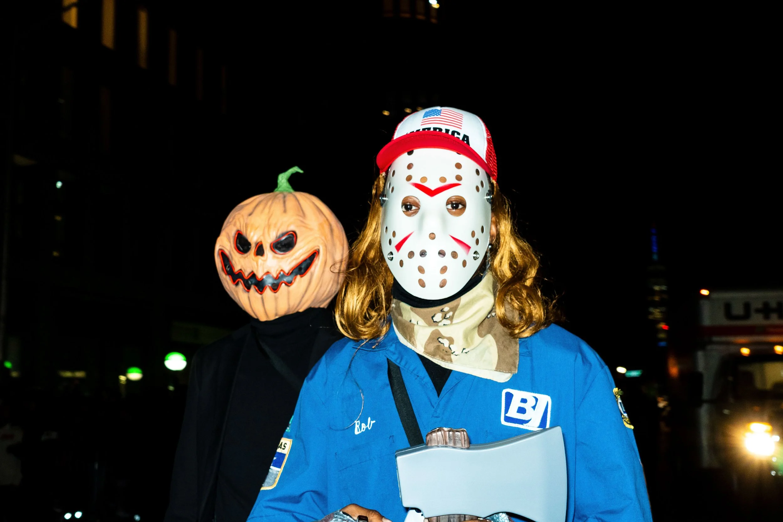 Two individuals wearing Halloween masks: one with a pumpkin head and a sinister face, the other with a white hockey-style mask with red markings, in a dark outdoor setting at night.