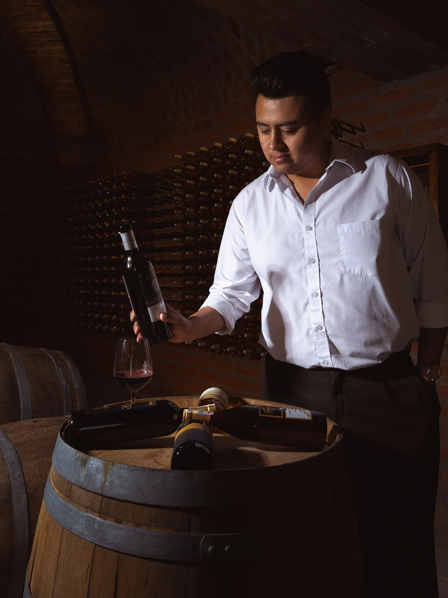 A man in a white shirt standing in a dimly lit wine cellar, holding a wine bottle, with wine bottles and glasses on a wooden barrel.