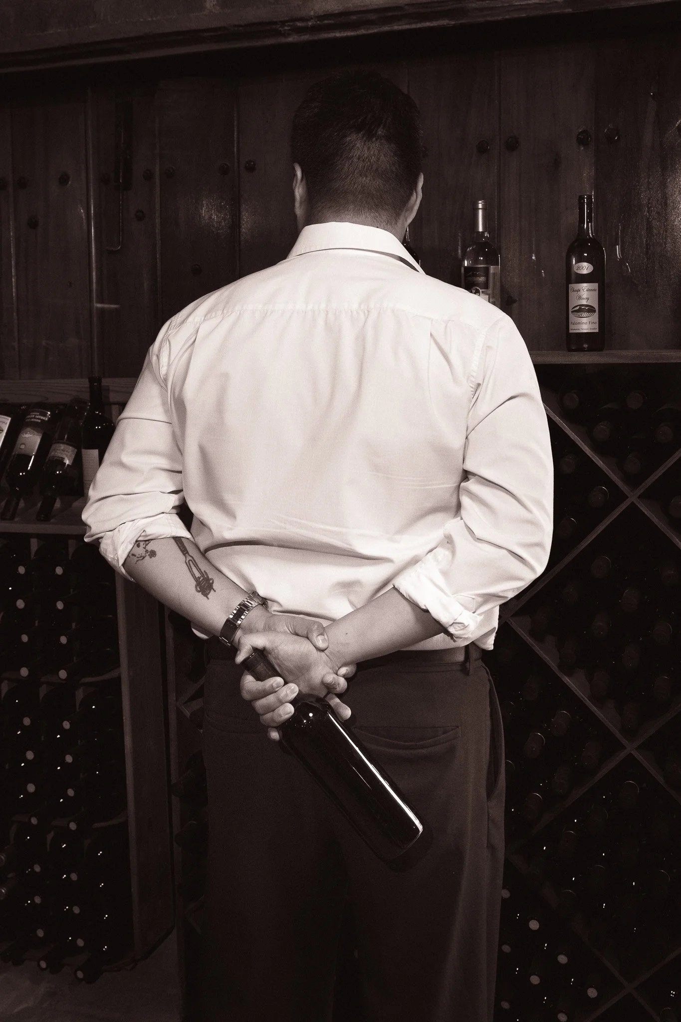 A man with short dark hair wearing a white dress shirt and black pants is standing with his back to the camera in a wine cellar or bar. He is holding a wine bottle behind his back with both hands. There are shelves of wine bottles on the dark wooden 