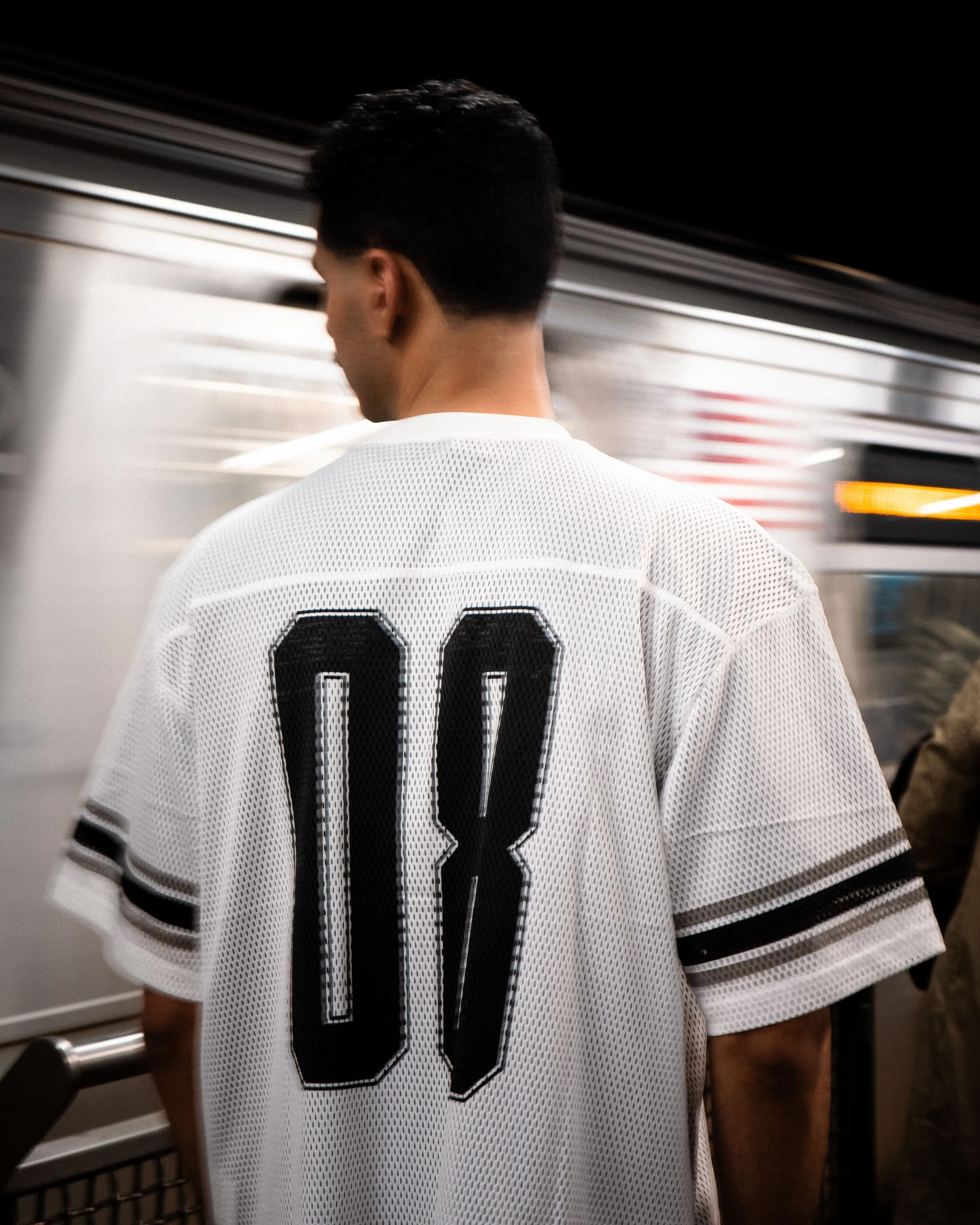 A person wearing a white sports jersey with the number 08 on the back, standing near a subway train.