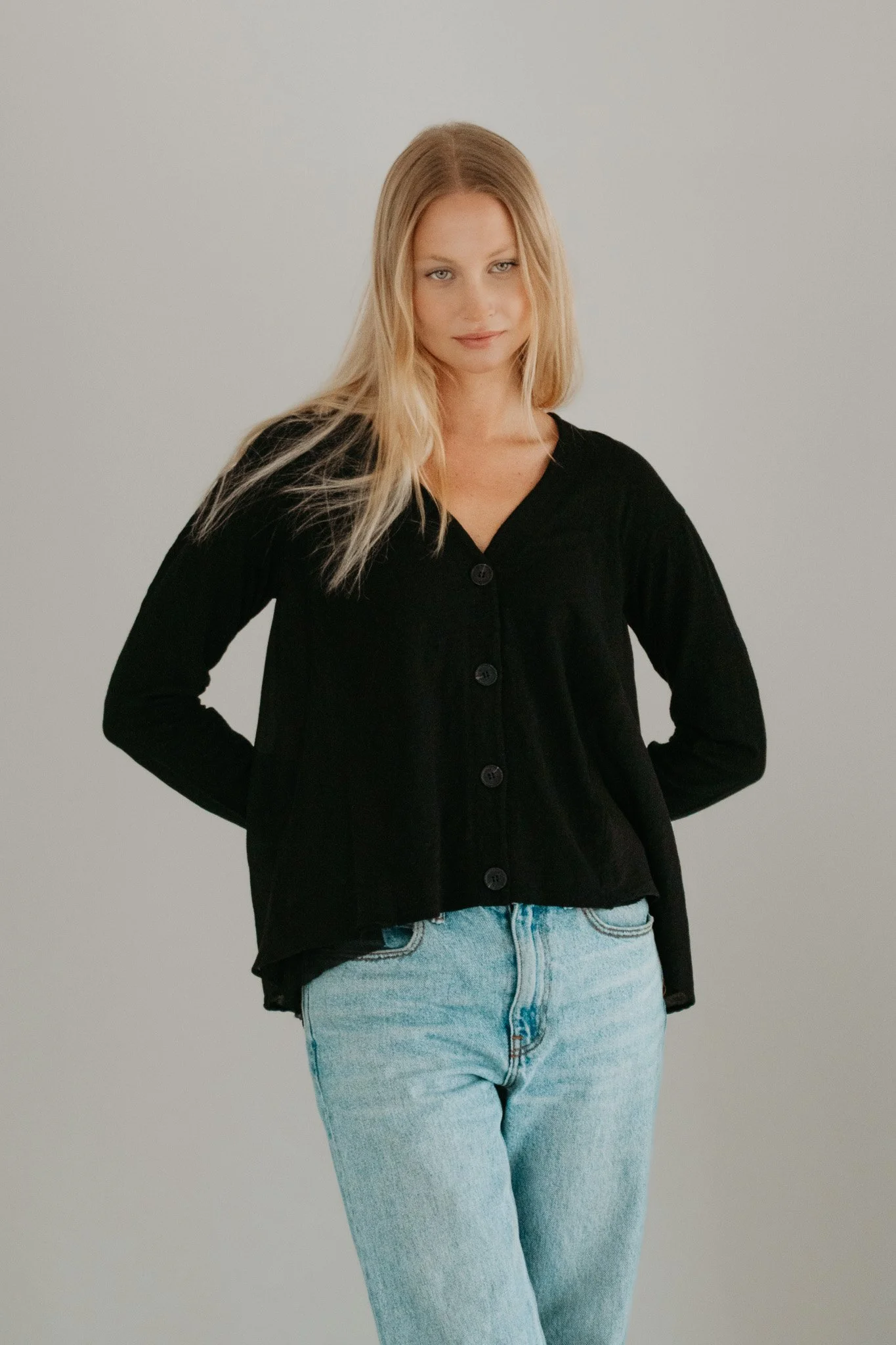 A young woman with long blonde hair, wearing a black cardigan and light blue jeans, standing against a plain white background.