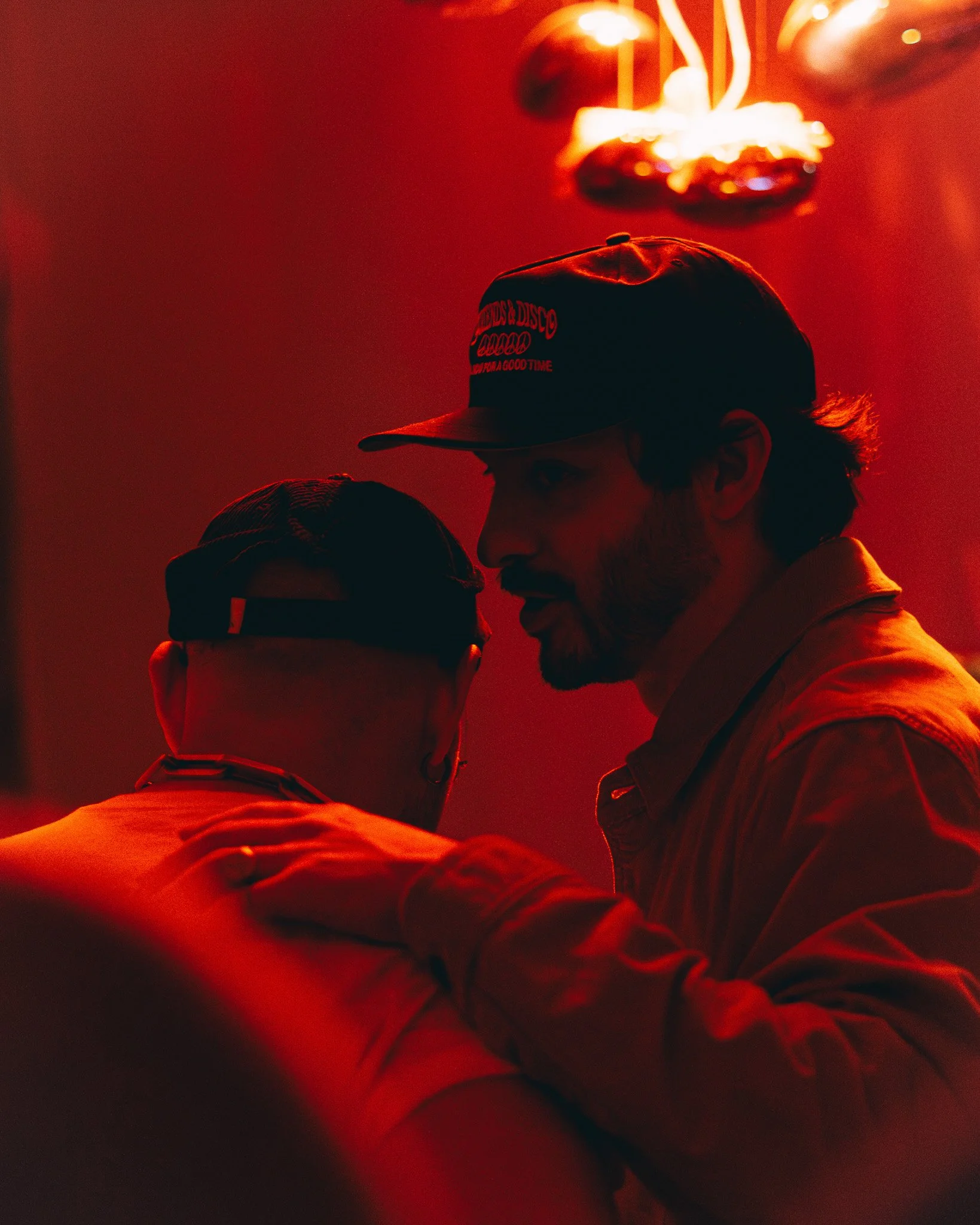 Two people, a man and a boy, are close together with their foreheads touching, in a dimly lit room illuminated by red light and a distinctive ceiling fan above them.