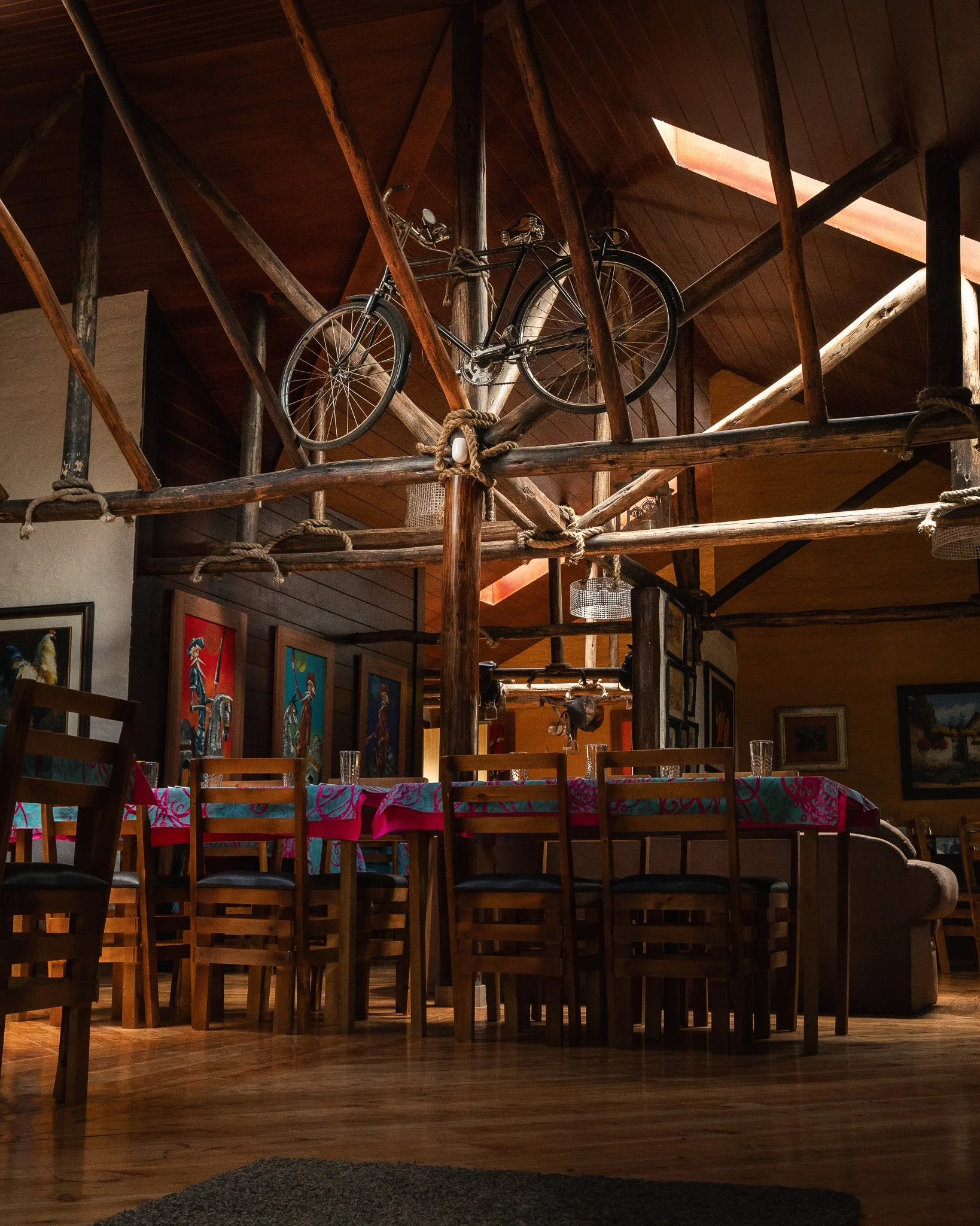 Interior of a cozy restaurant or café with wooden accents. There is a dining table with a pink and blue patterned tablecloth, surrounded by wooden chairs. The upper level features a rustic wooden railing with a bicycle mounted on it for decoration. A