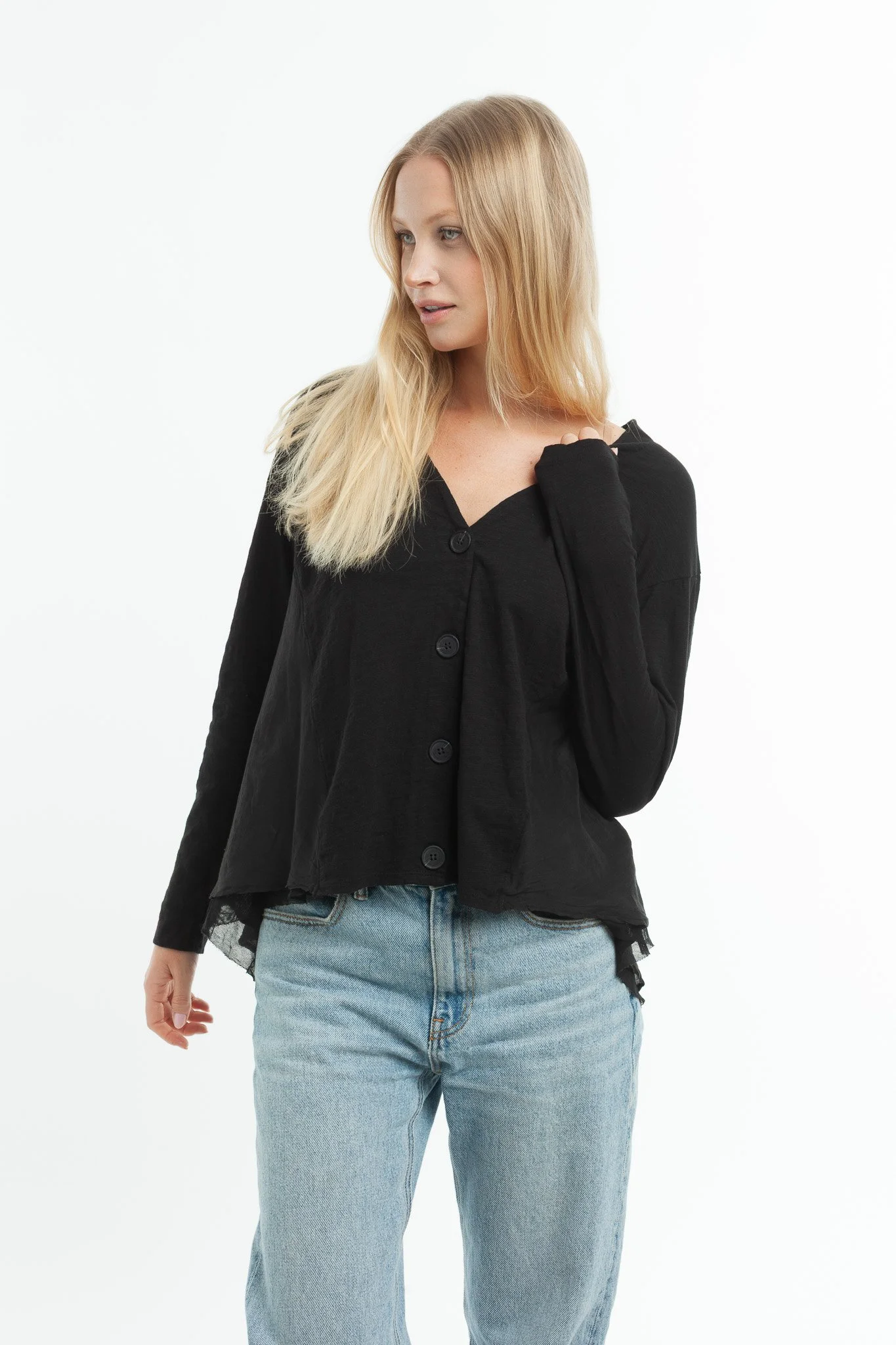 A woman with long blonde hair wearing a black button-up shirt and light blue jeans standing against a white background.