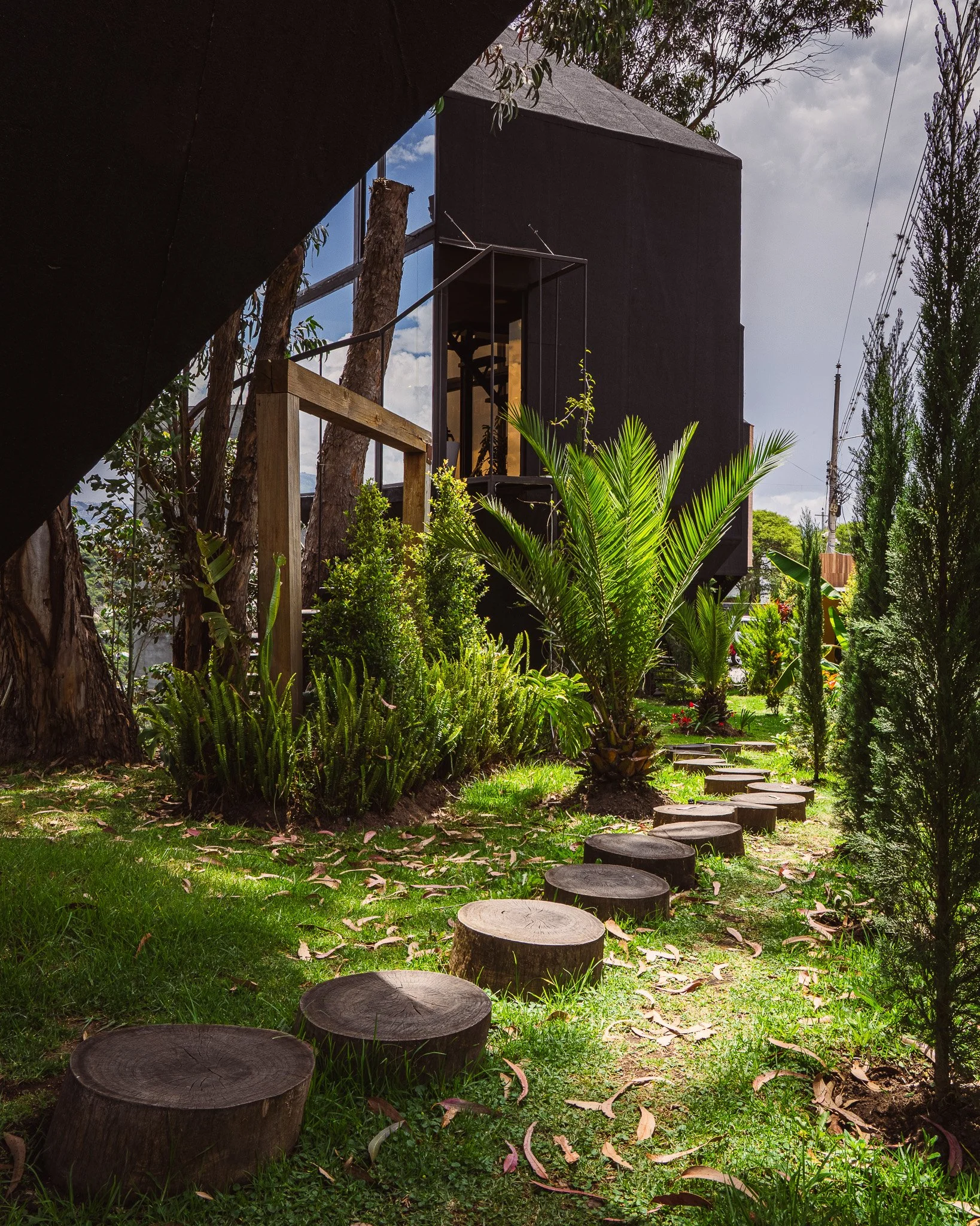 Step stones in a grassy garden with lush green plants, trees, and a modern black building with glass windows in the background.
