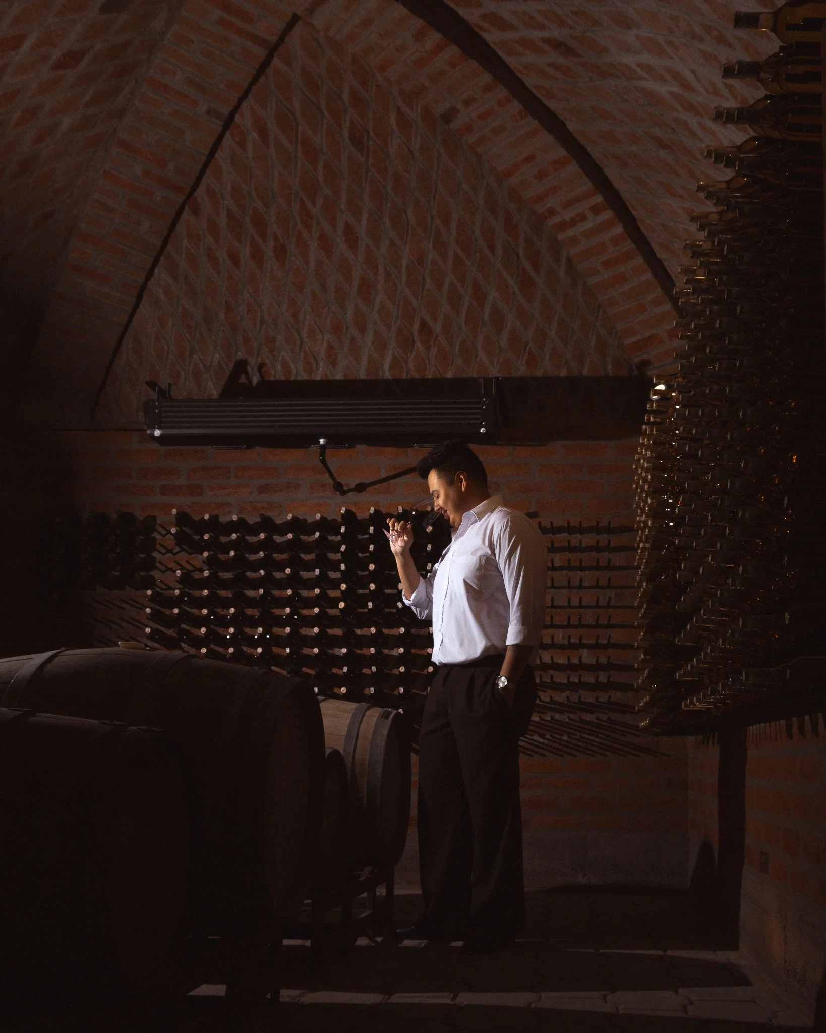 A man in a white shirt standing in front of a wine rack in a room with a brick wall ceiling.