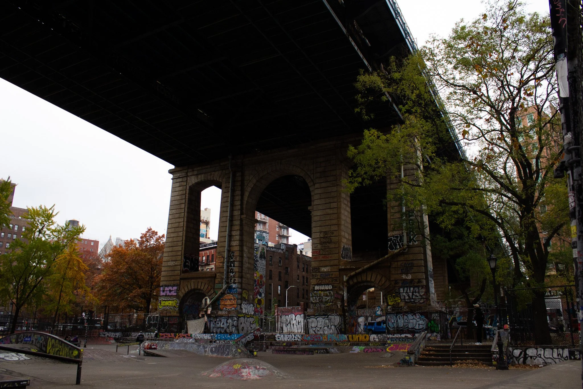 Underneath an elevated bridge with graffiti-covered supports, a skate park with ramps and a fence in an urban area. There are trees with green and orange foliage around.
