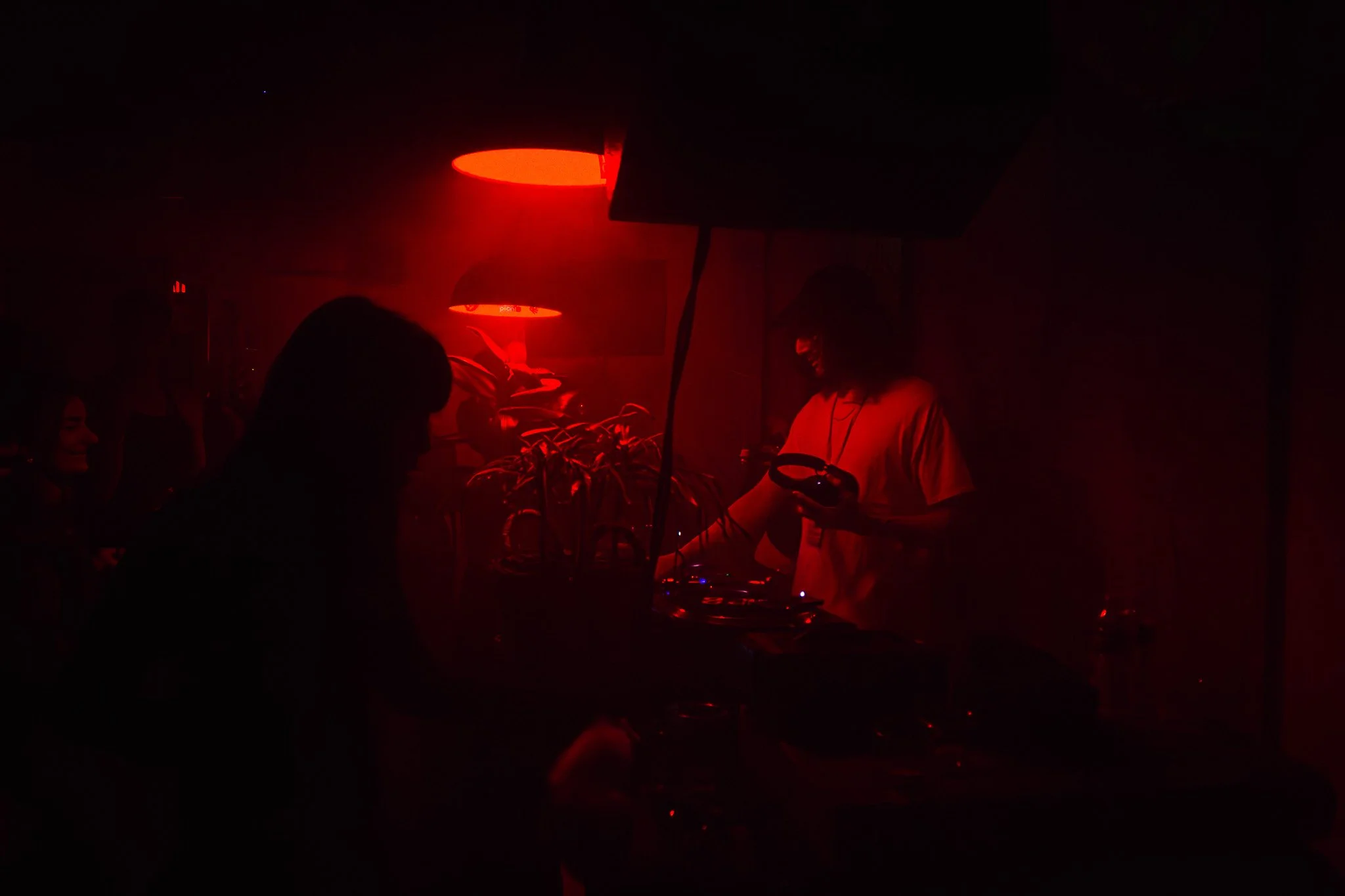 People at a dimly lit indoor party or club with red lighting, some silhouettes visible, one person is using a phone, and a DJ setup or audio equipment is present.