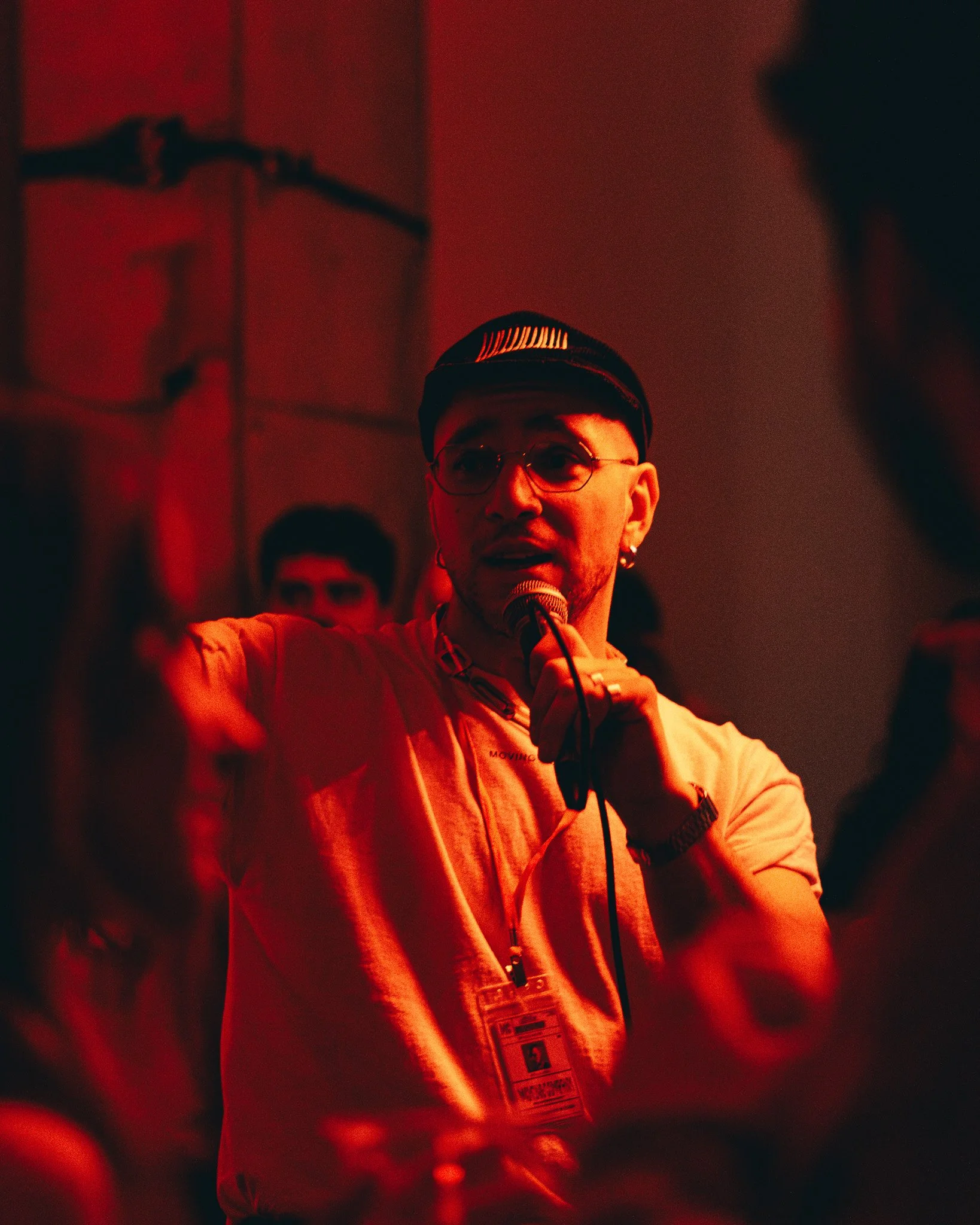 A person with glasses and earrings holding a microphone and wearing a hoodie, illuminated by red lighting, with a dark background.