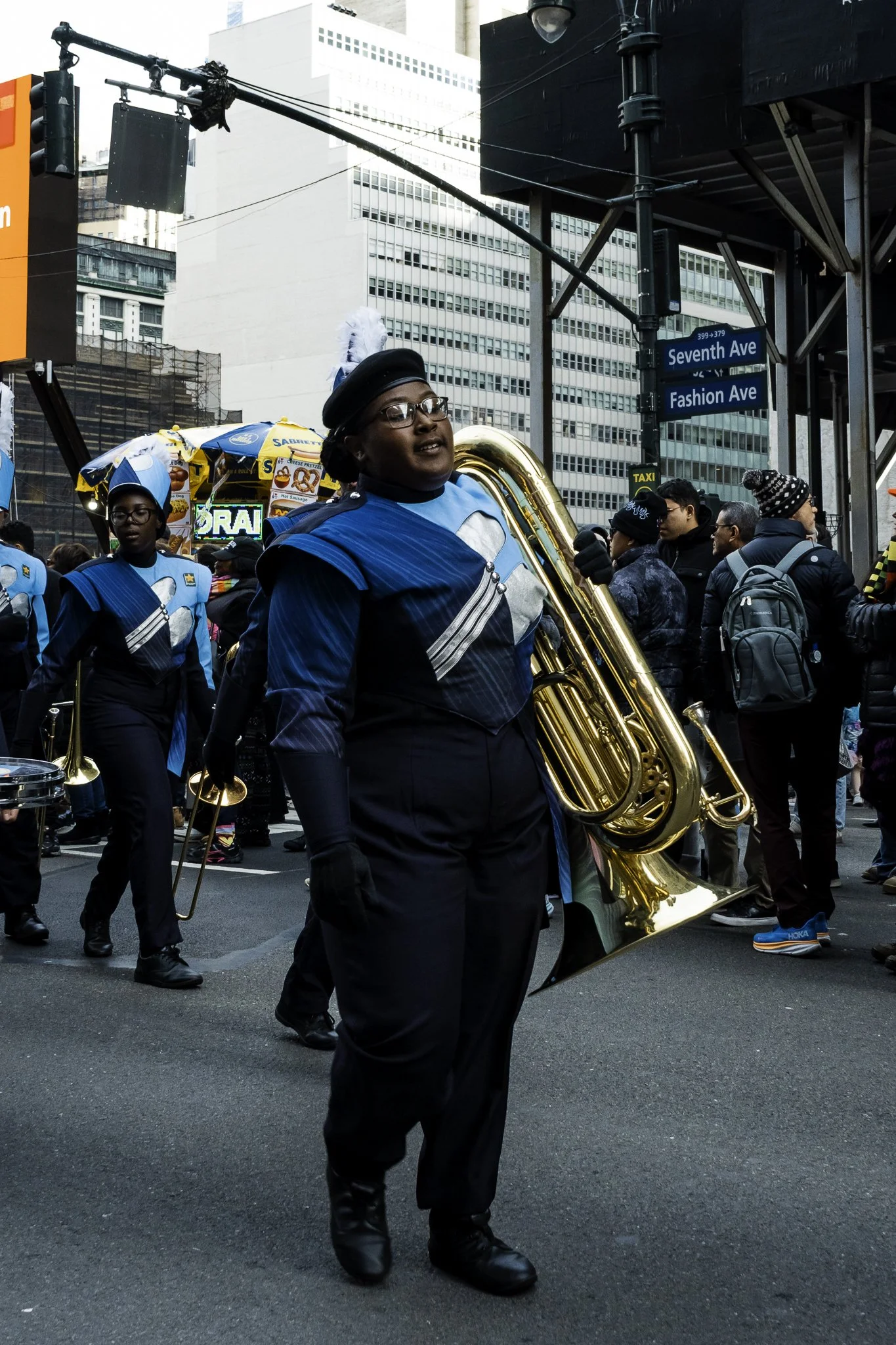 Marching band members in matching blue and black uniforms walking down a city street with tall buildings, traffic lights, and street signs for Seventh Ave and Fashion Ave.