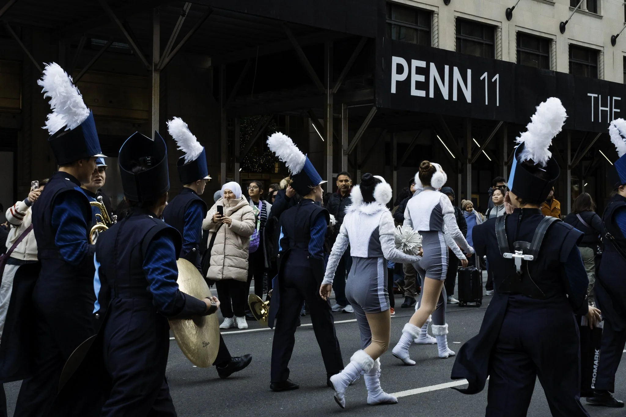 A street parade with musicians in black uniforms and feathered hats, and a woman in reindeer costume, in front of a building with a sign reading 'PENN 11'.