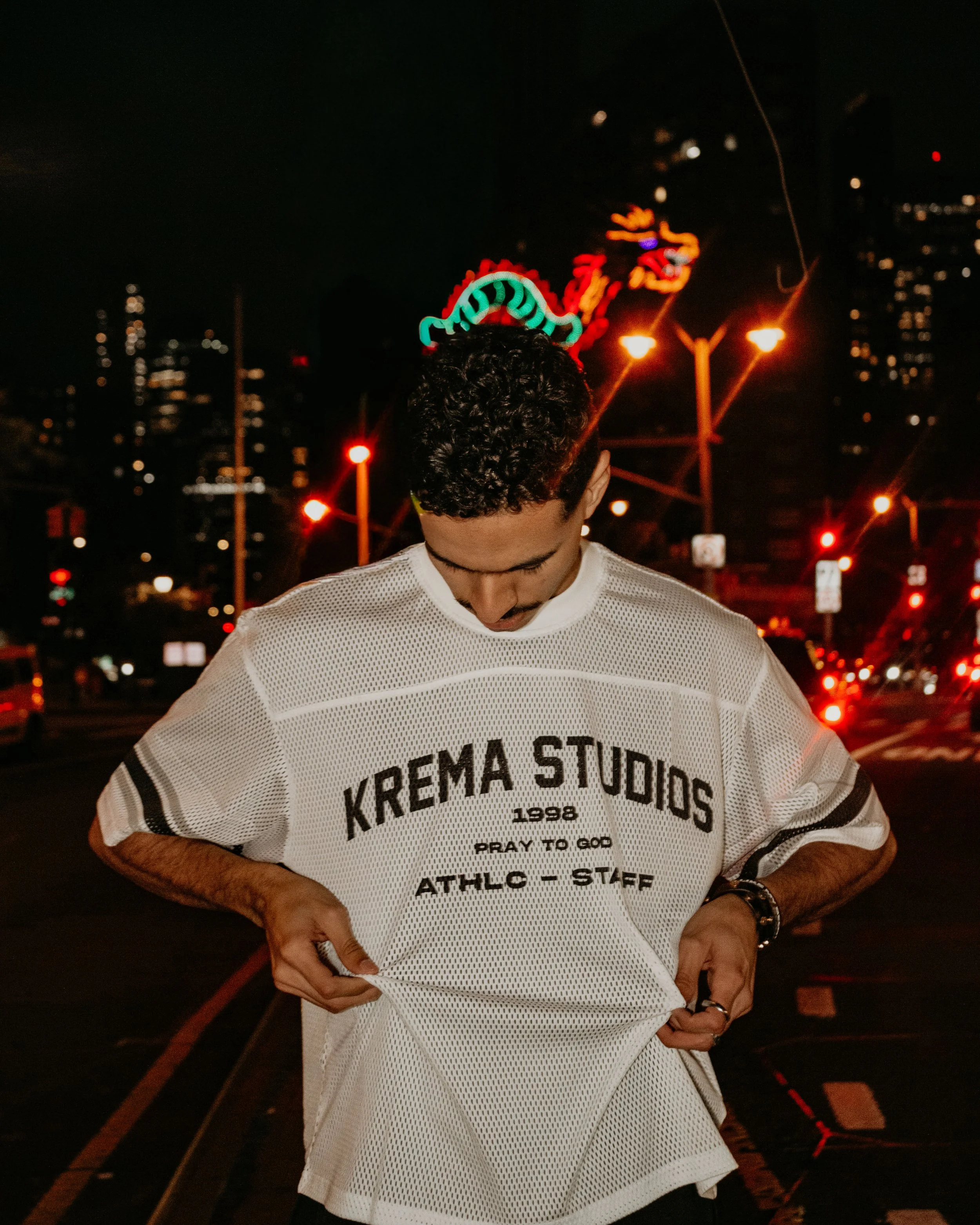 A man with curly hair looking down, wearing a white sports jersey with black text that reads 'KREMA STUDIOS 1998 PRAY TO GOD ATHLO - STAFF', standing in a city street at night with bright neon lights, a dragon sign in the background, and traffic ligh