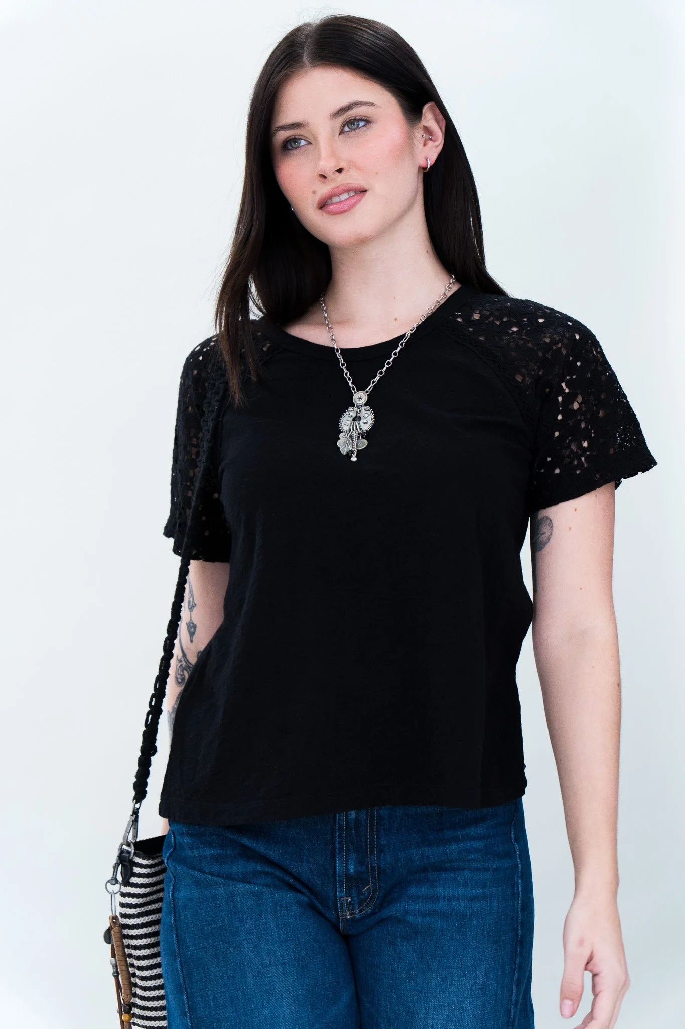 A young woman with dark brown hair, blue eyes, and fair skin wearing a black lace short-sleeve top, blue jeans, and silver jewelry, standing against a plain white background.