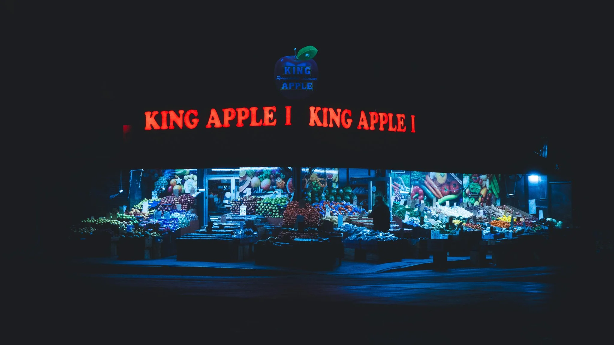 Night view of a fruit market stall with various fresh produce, illuminated by bright lights. A large neon sign reads 'KING APPLE' in red letters, with additional signage displaying fruit images and logos.