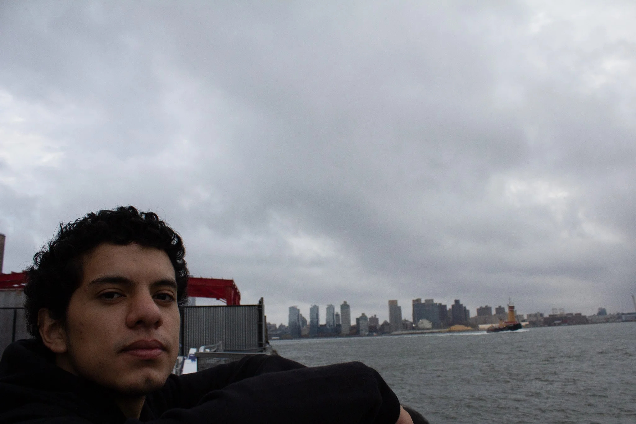 Young man with dark curly hair and a black jacket, sitting by the water with a city skyline of tall buildings and a cloudy sky in the background.