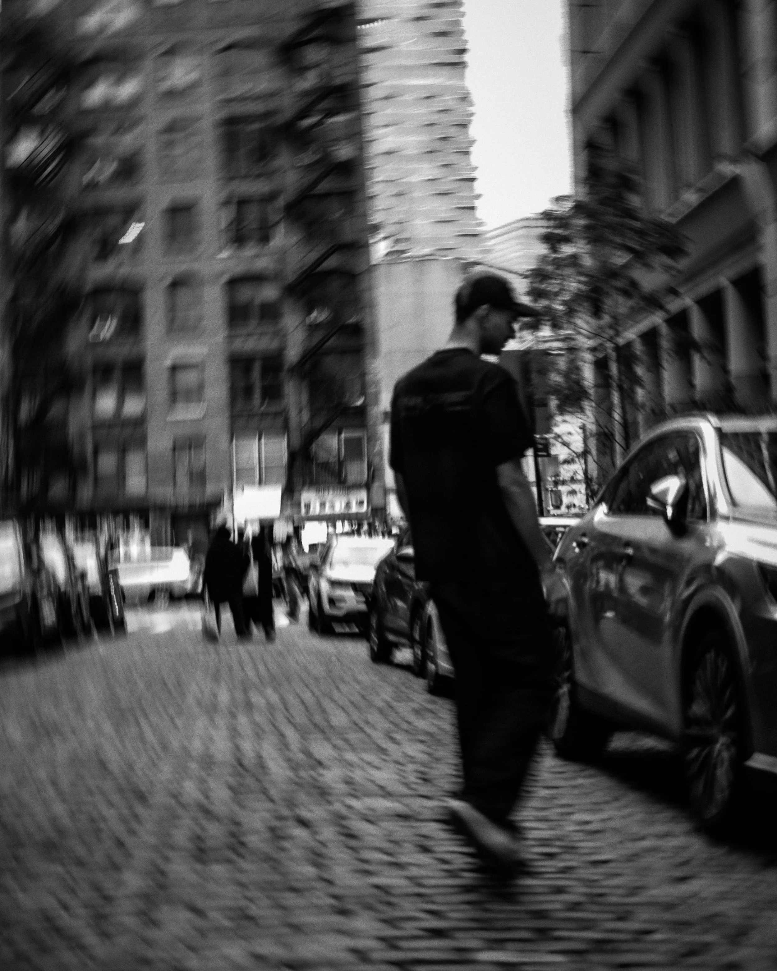 A black and white photo of a city street with a man in the foreground looking at his phone or device. Several cars are parked along the curb, and buildings with multiple windows are visible in the background.