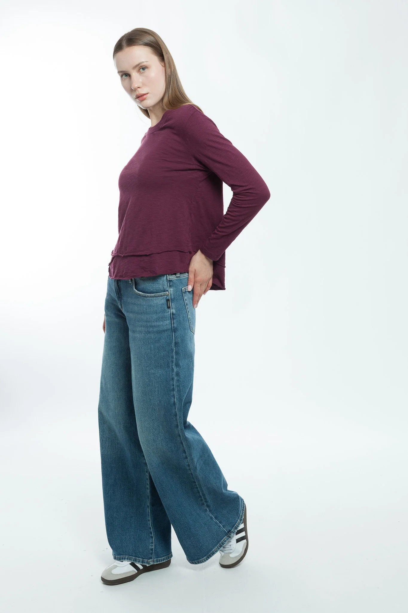 A young woman with light brown hair and blue eyes wearing a maroon long-sleeve shirt, wide-leg blue jeans, and white sneakers, posing against a plain white background.