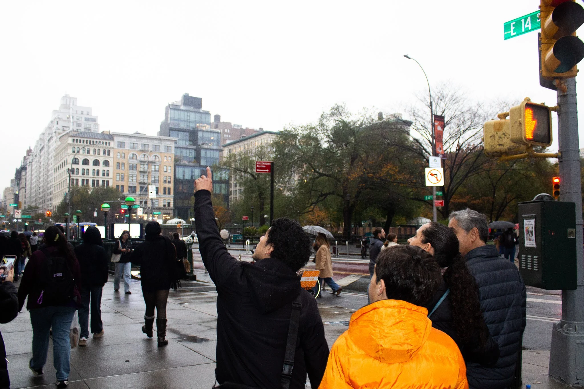 Group of people waiting at a crosswalk on a city street in rainy weather with trees and tall buildings in the background.