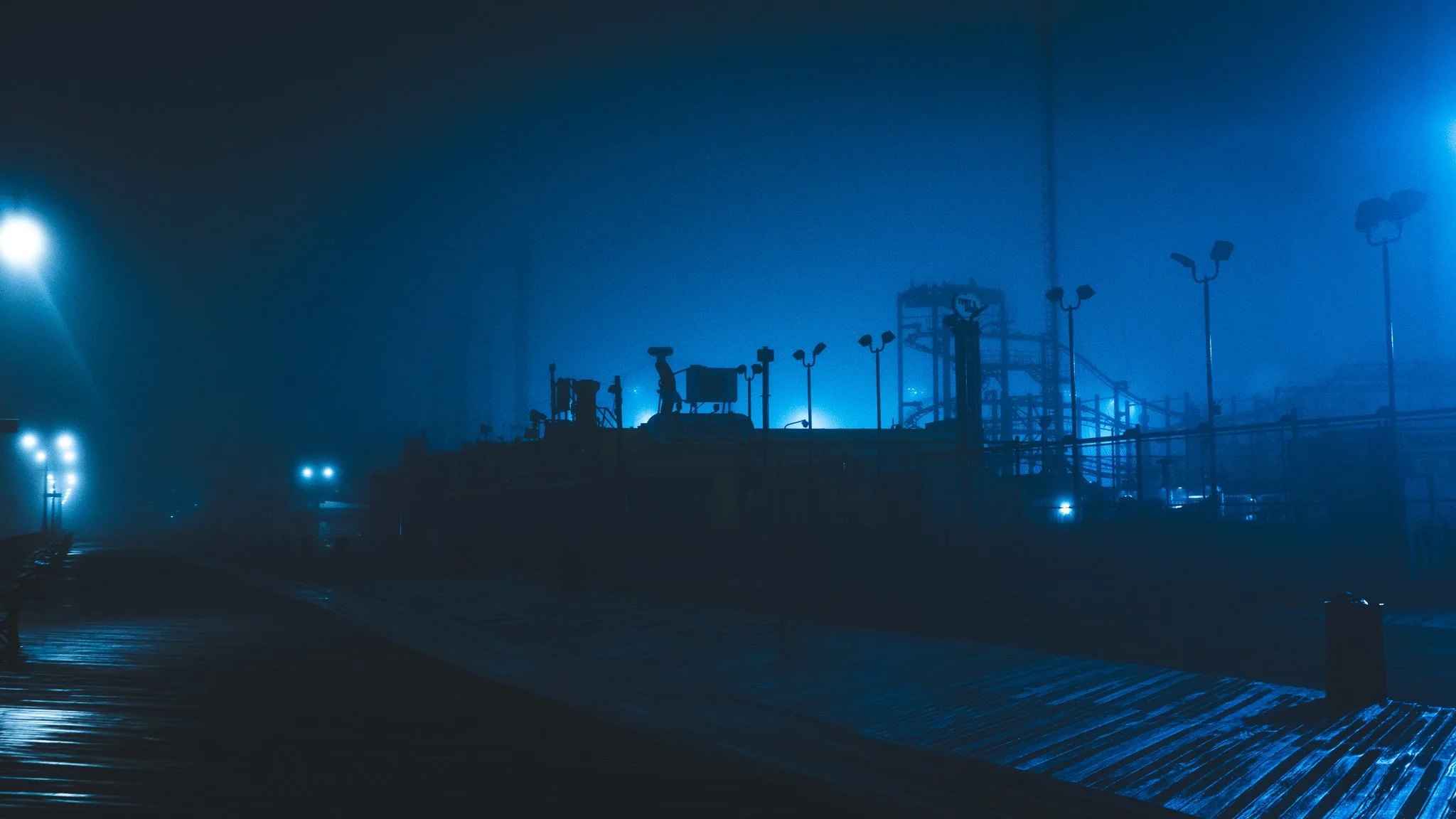 Night scene of an industrial facility or dock shrouded in fog, with bright blue lighting illuminating the structures and walkways.