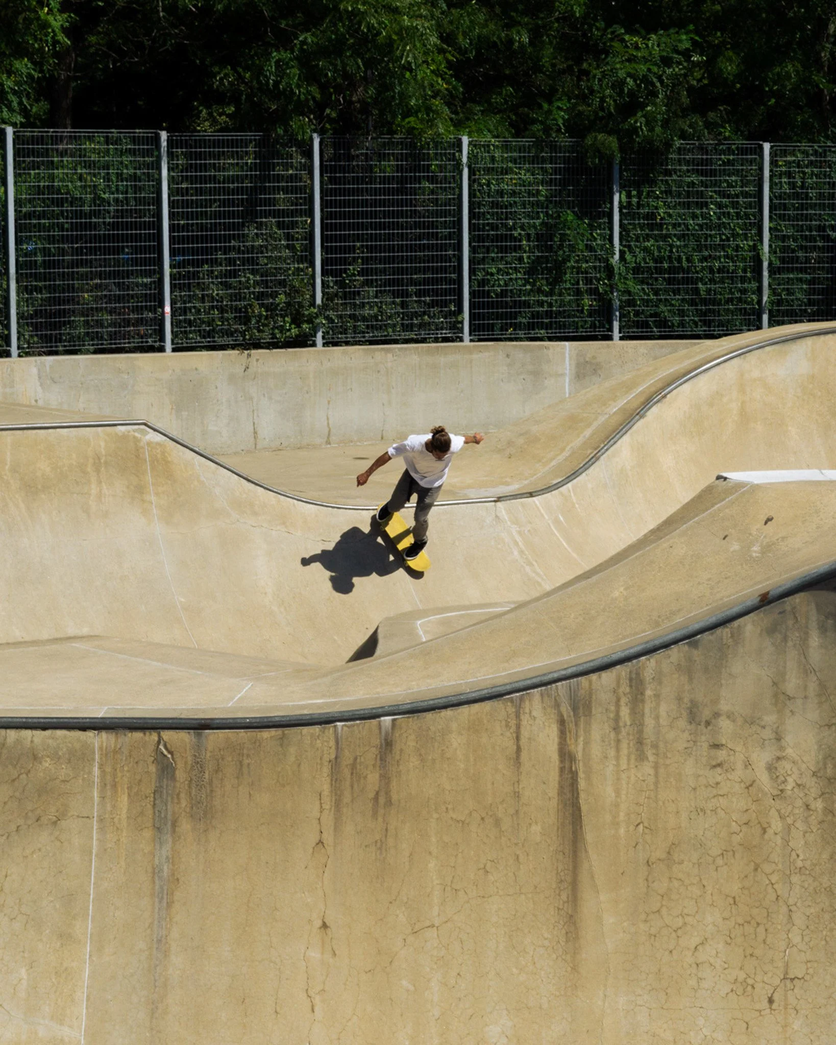 A skateboarder wearing a white shirt and dark pants riding in a concrete skatepark with a yellow skateboard.