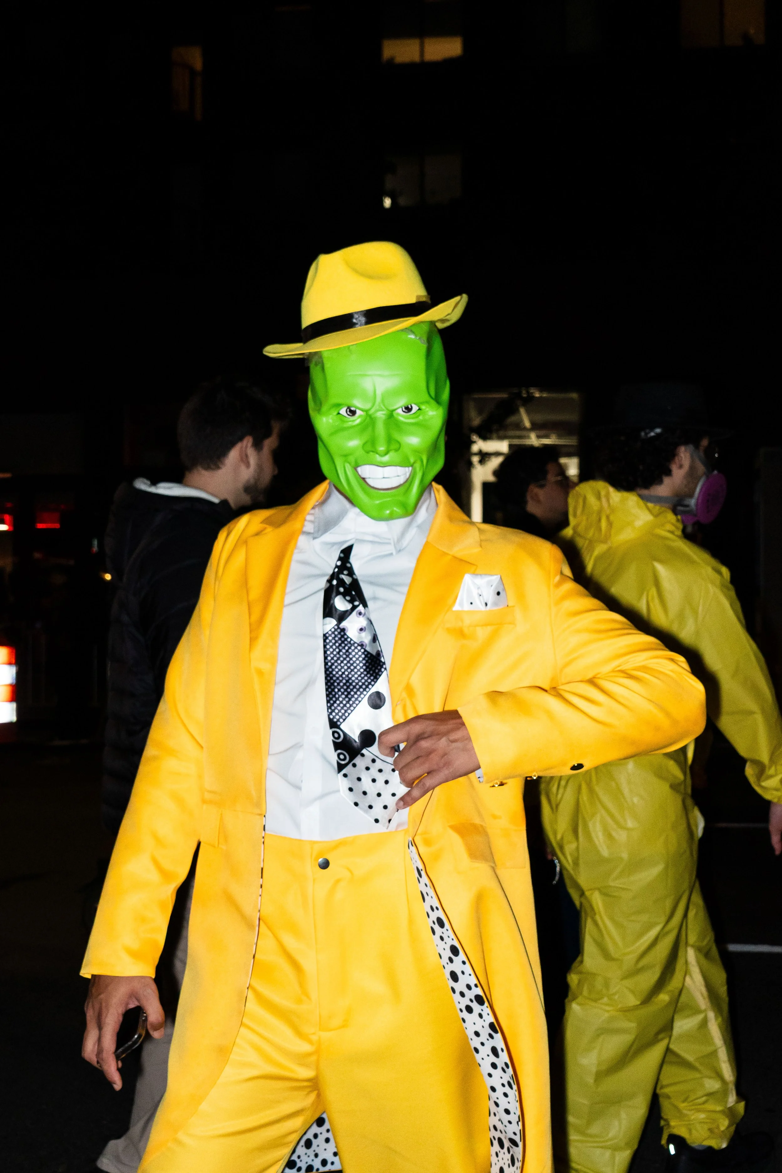 Person in a bright yellow suit with a matching hat, wearing a green mask resembling a comic book character, at a nighttime event.
