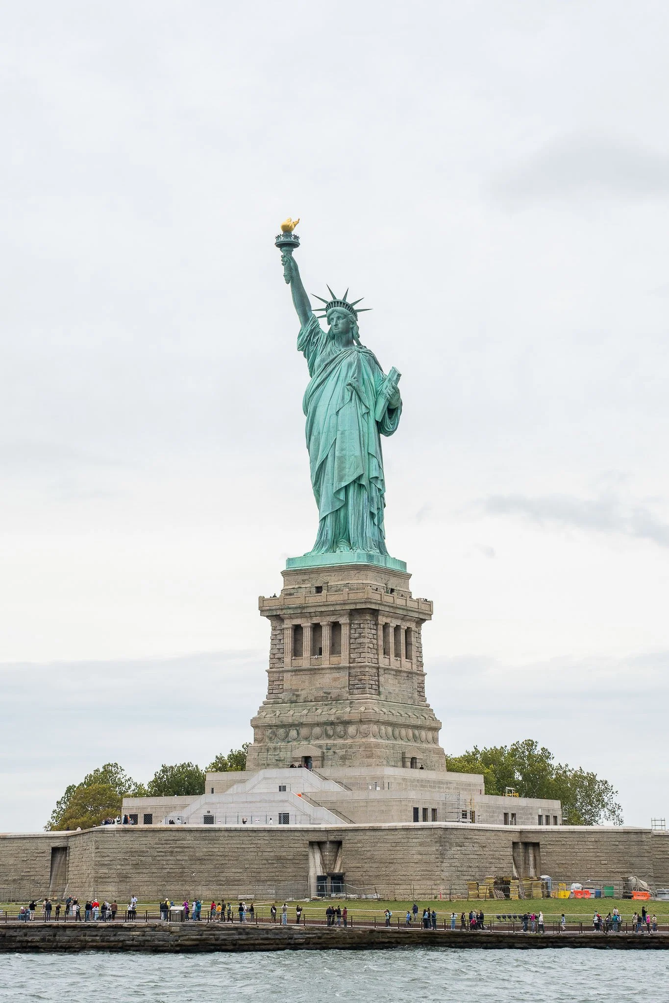 The Statue of Liberty standing on a pedestal by the water, with people walking along the waterfront.