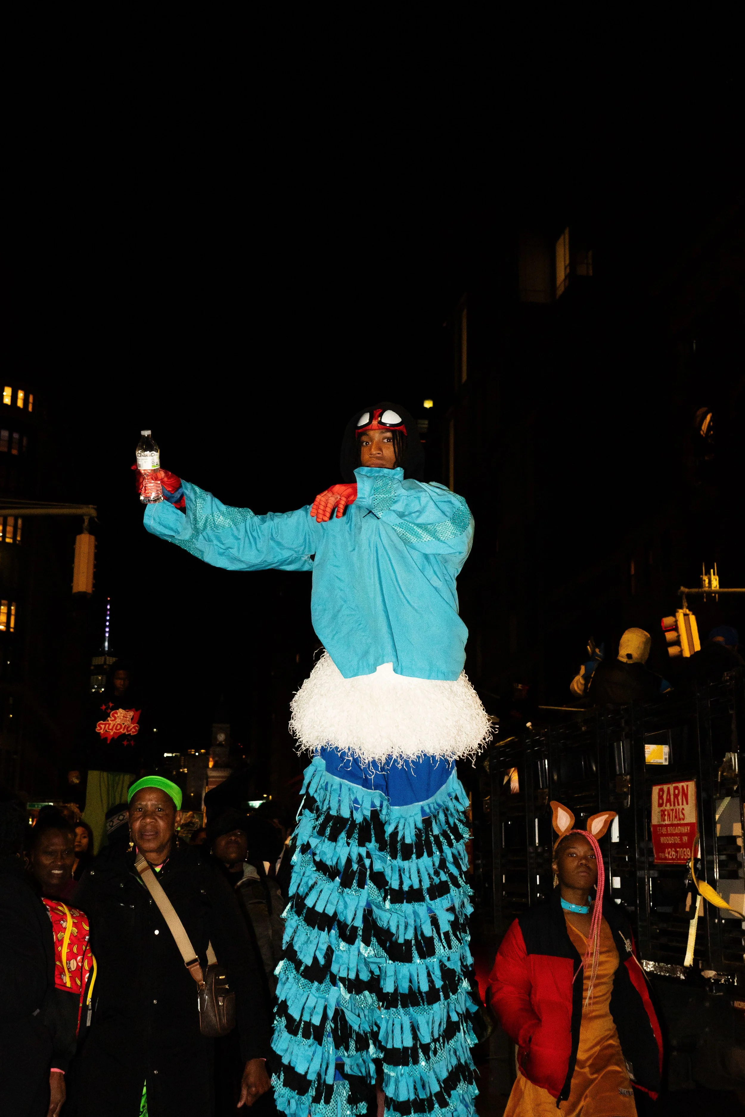 Person in a blue jacket and colorful costume standing on stilts at a night street event, with others around, holding a water bottle, some wearing costumes and headpieces.