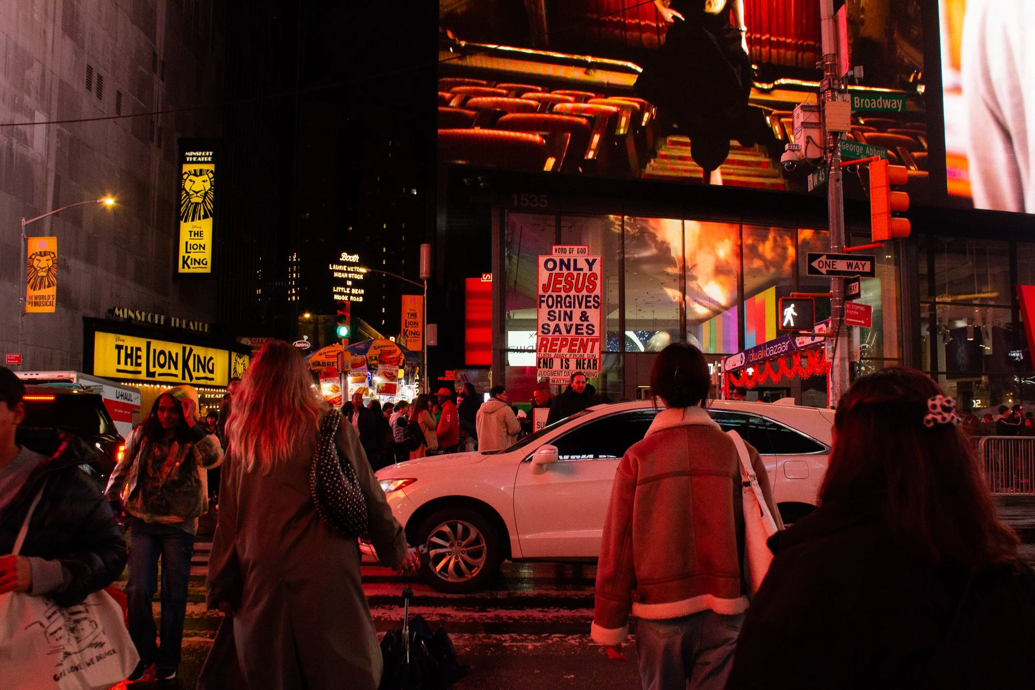 Nighttime scene in Times Square, New York City, with people walking on the street, bright red lights, billboards, and signs including 'The Lion King' and religious posters.