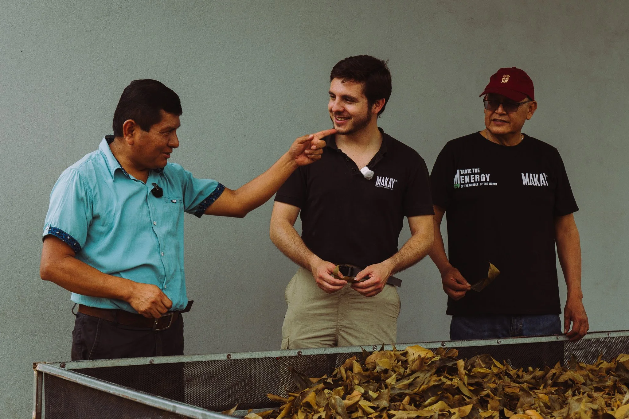 Three men standing behind a table of dried leaves. The man on the left is wearing a light blue shirt, the man in the middle is in a black shirt with a microphone clipped to it, and the man on the right is wearing a black shirt and a red cap. The man 
