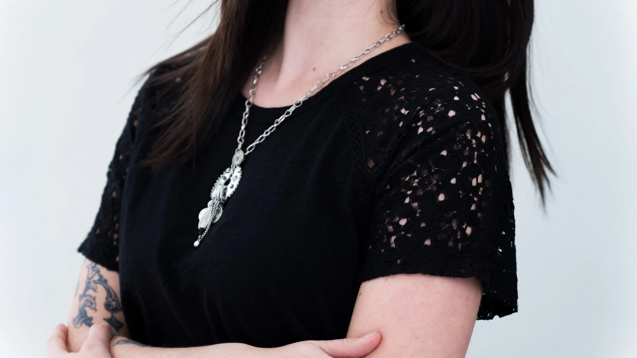 Close-up of a woman wearing a black top with lace sleeves and a silver chain necklace with pendants, displaying tattoos on her forearm and hand.