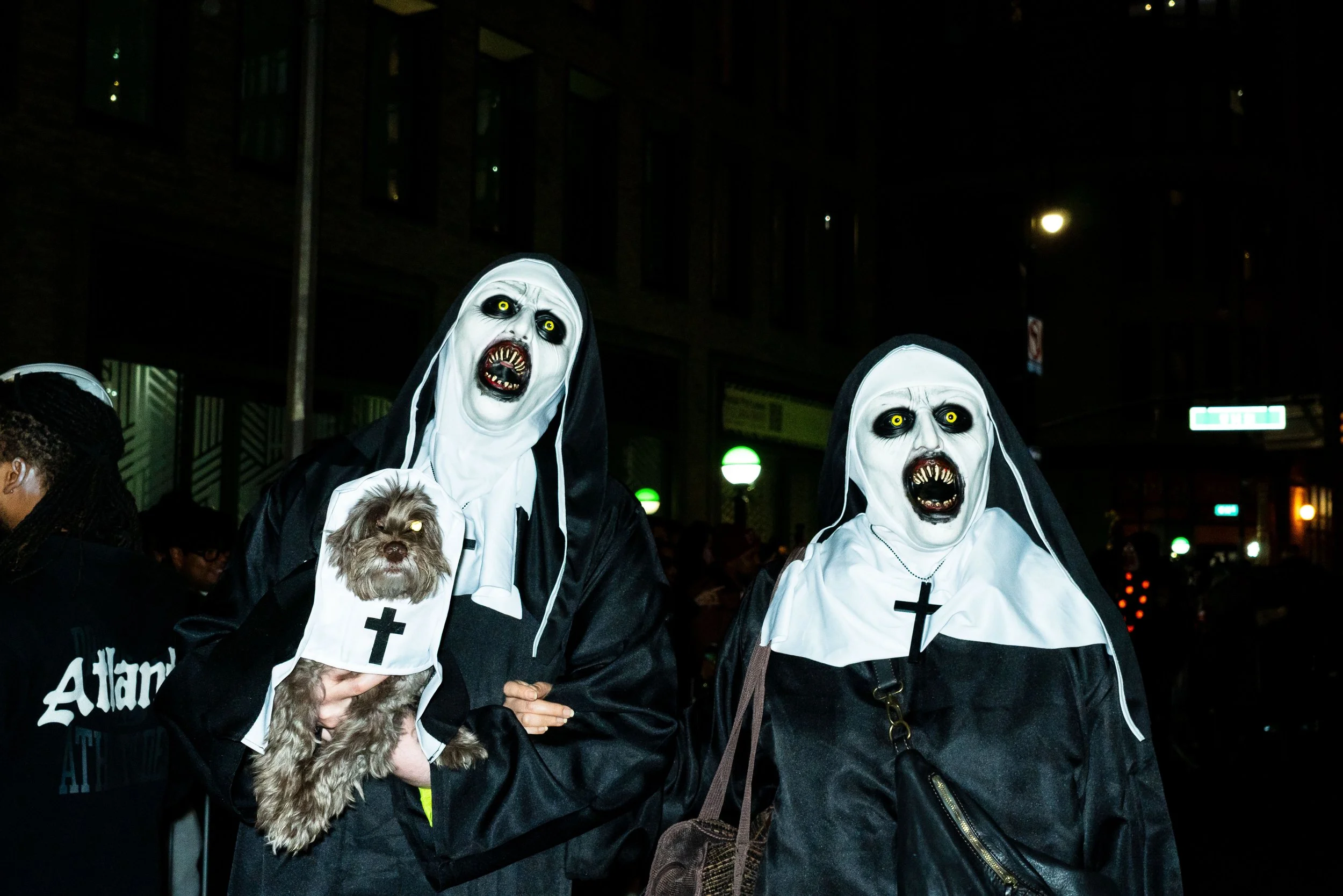 Two people dressed as scary nuns in black and white habits with zombie faces, open mouths, and yellow eyes, walking at night in a city street, holding a small dog in a nun costume.