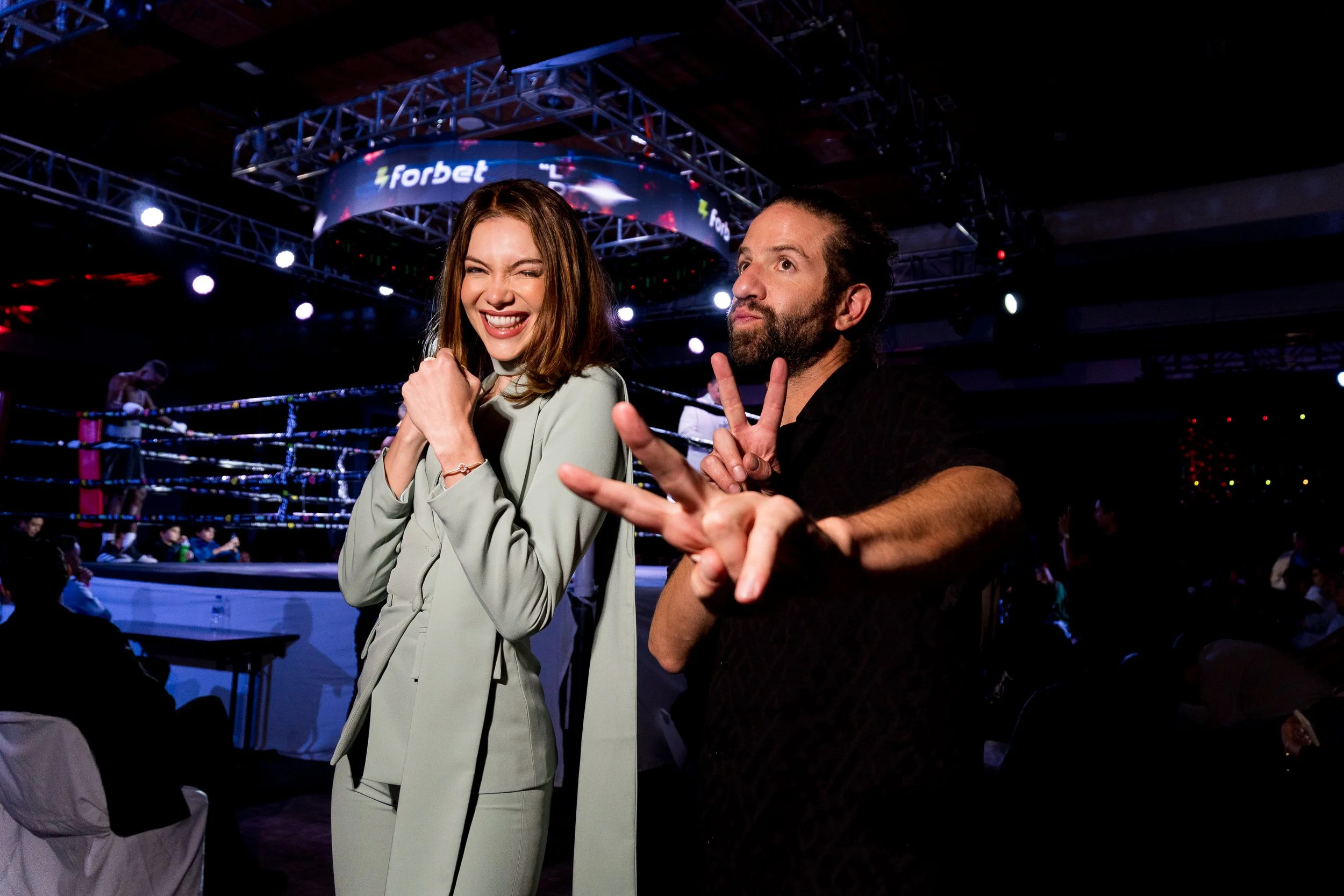 Two people posing in a boxing ring, with one person winking and the other making a peace sign, under a large digital screen showing the word 'Forbet'.
